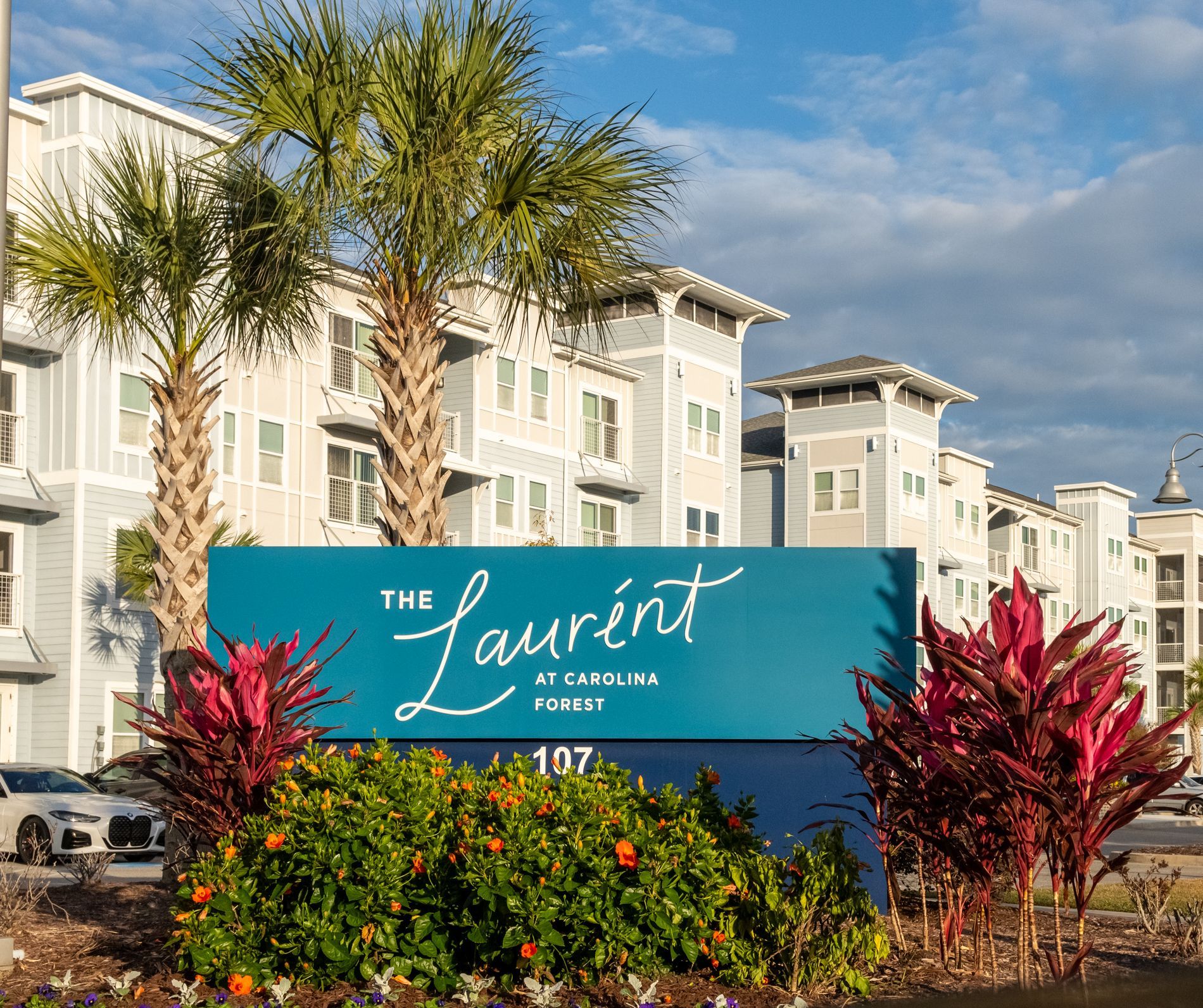 The Laurent apartment complex monument sign with teal background. Palm trees, and flowers surrounded in front of buildings.