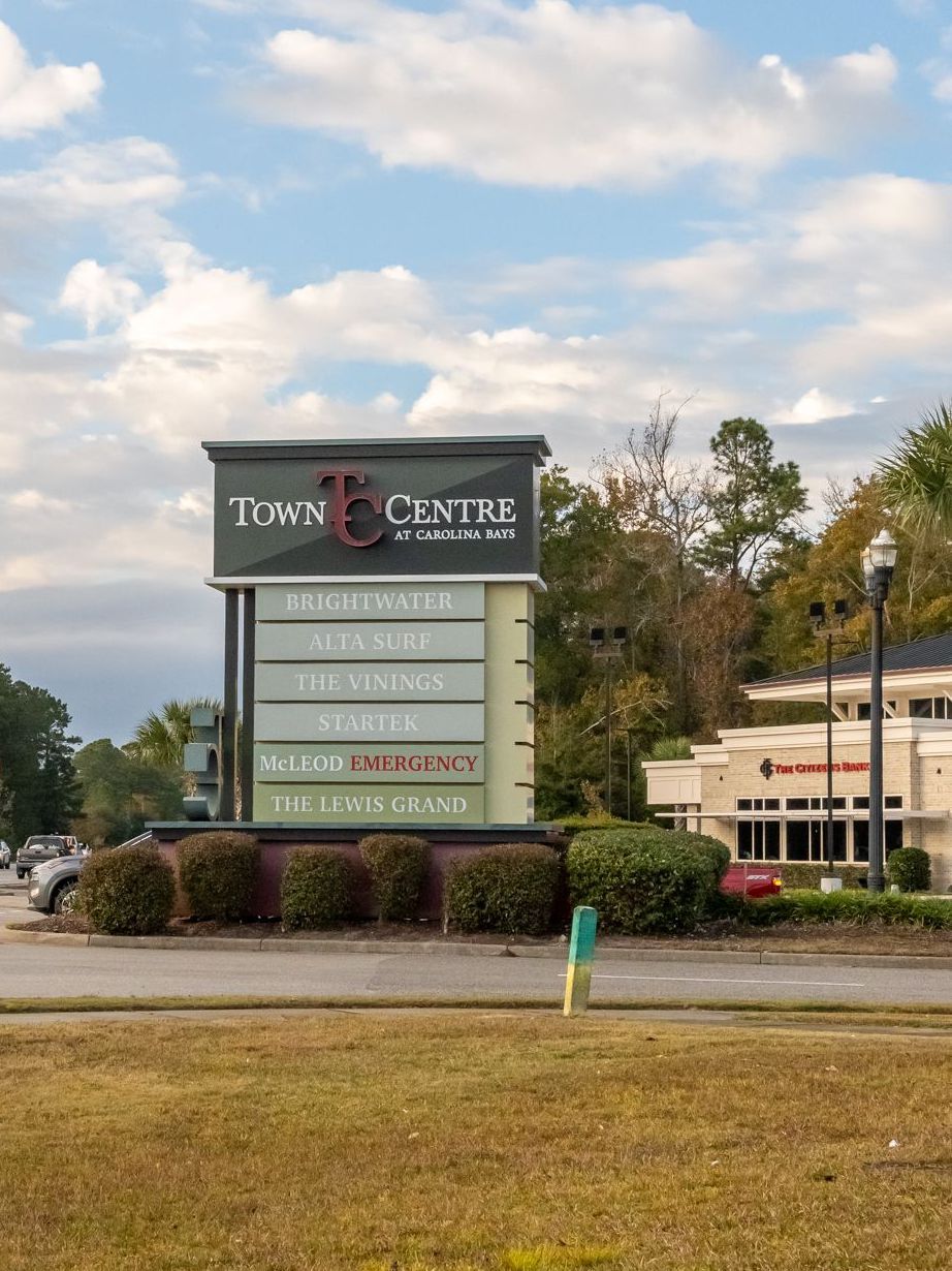 Town Centre sign with tenant listings, shrubs in front, building and cars in background, daytime.