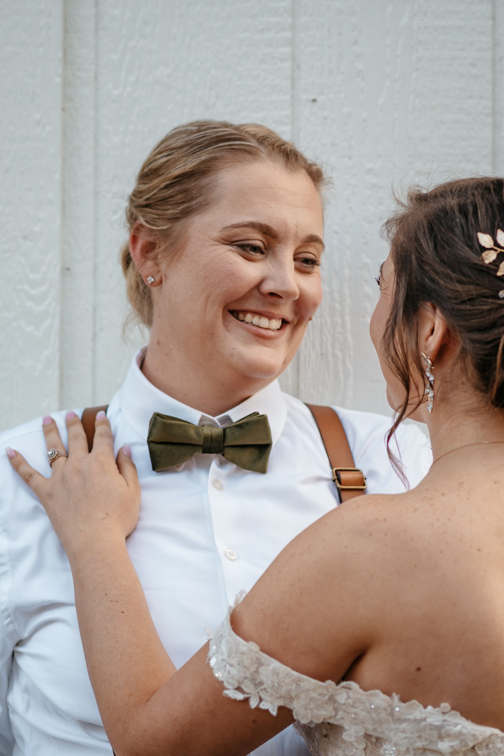 A bride and groom are looking at each other and smiling . the bride is wearing a bow tie and suspenders.