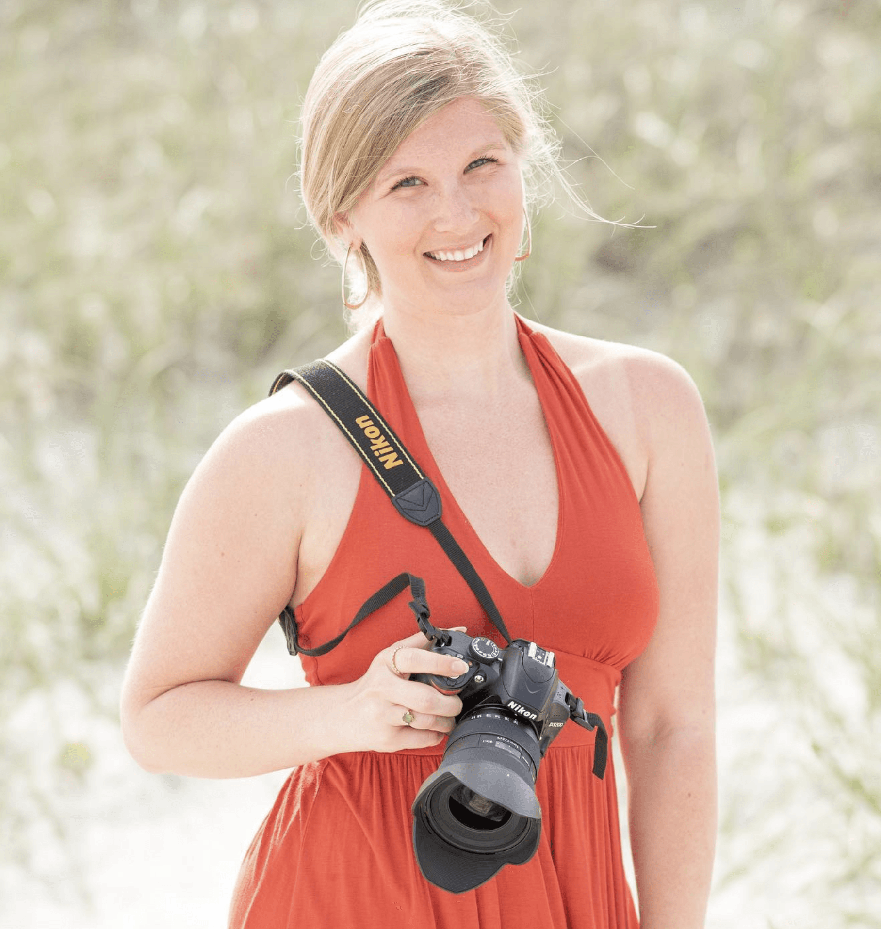 A woman, Morgan René Doscher (owner of Mo Knows Marketing, LLC DBA Mo Knows Art) in an orange dress is holding a nikon camera