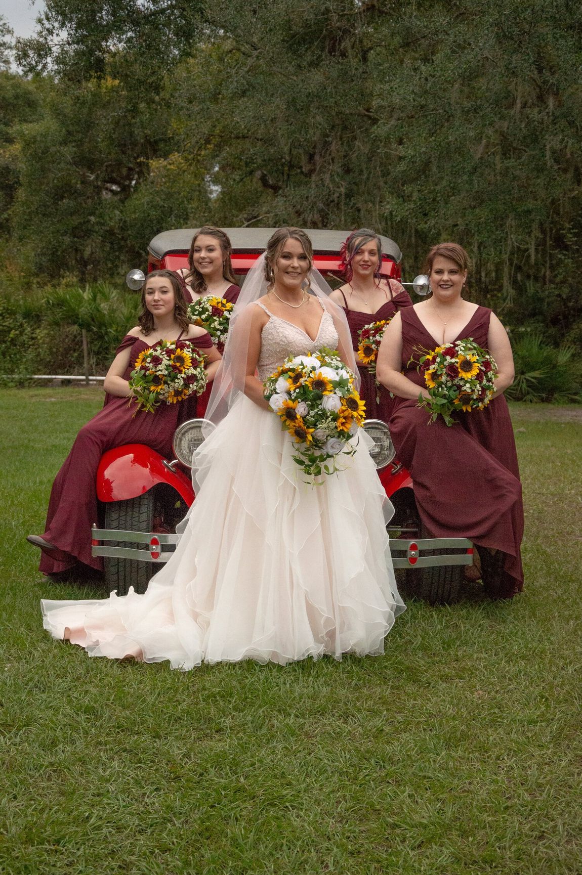 A bride and her bridesmaids are posing for a picture in front of an old red model t car holding sunflower bouquets.