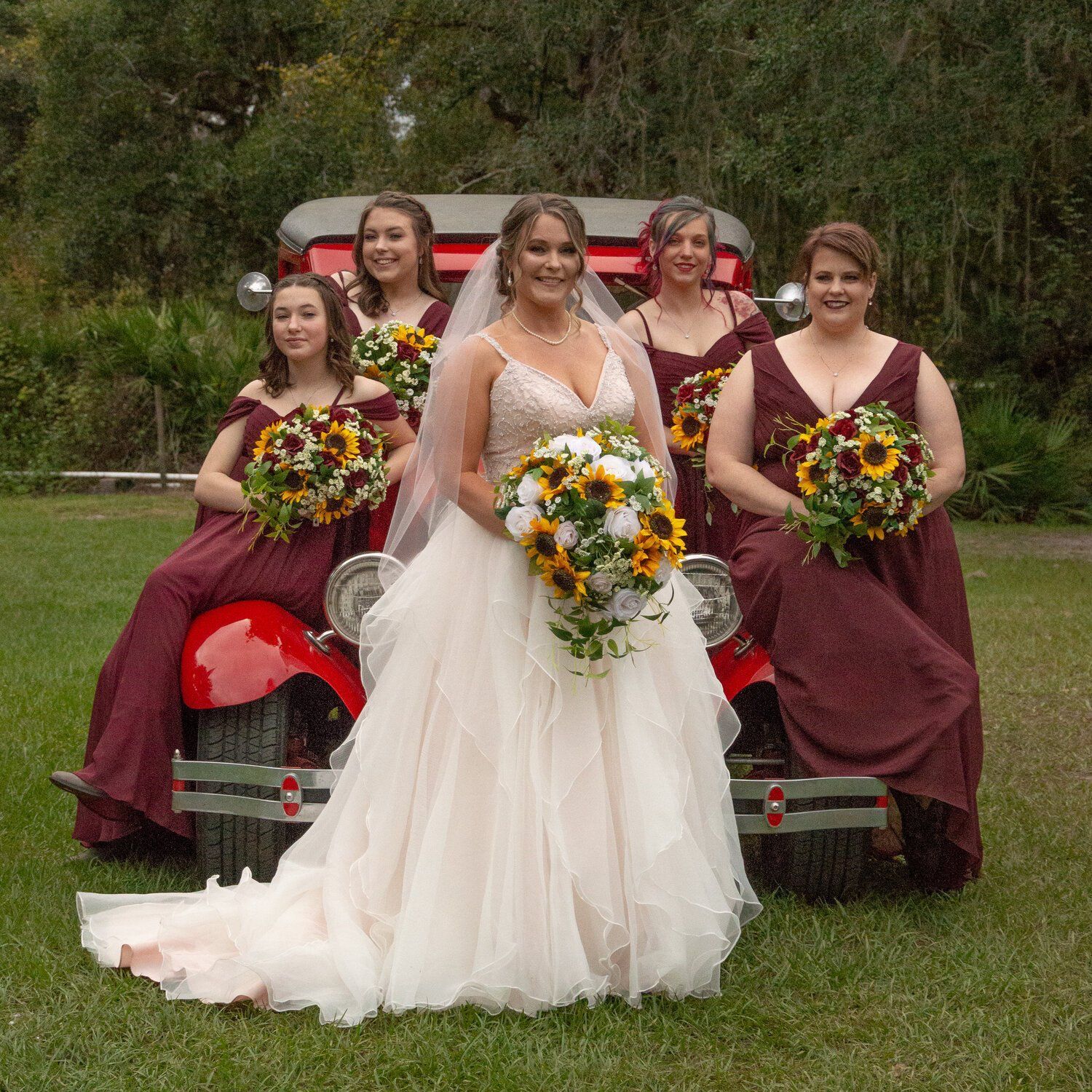 A group of bridesmaids on a old red car with the bride in the middle holding sunflower bouquets photo by Morgan Doscher