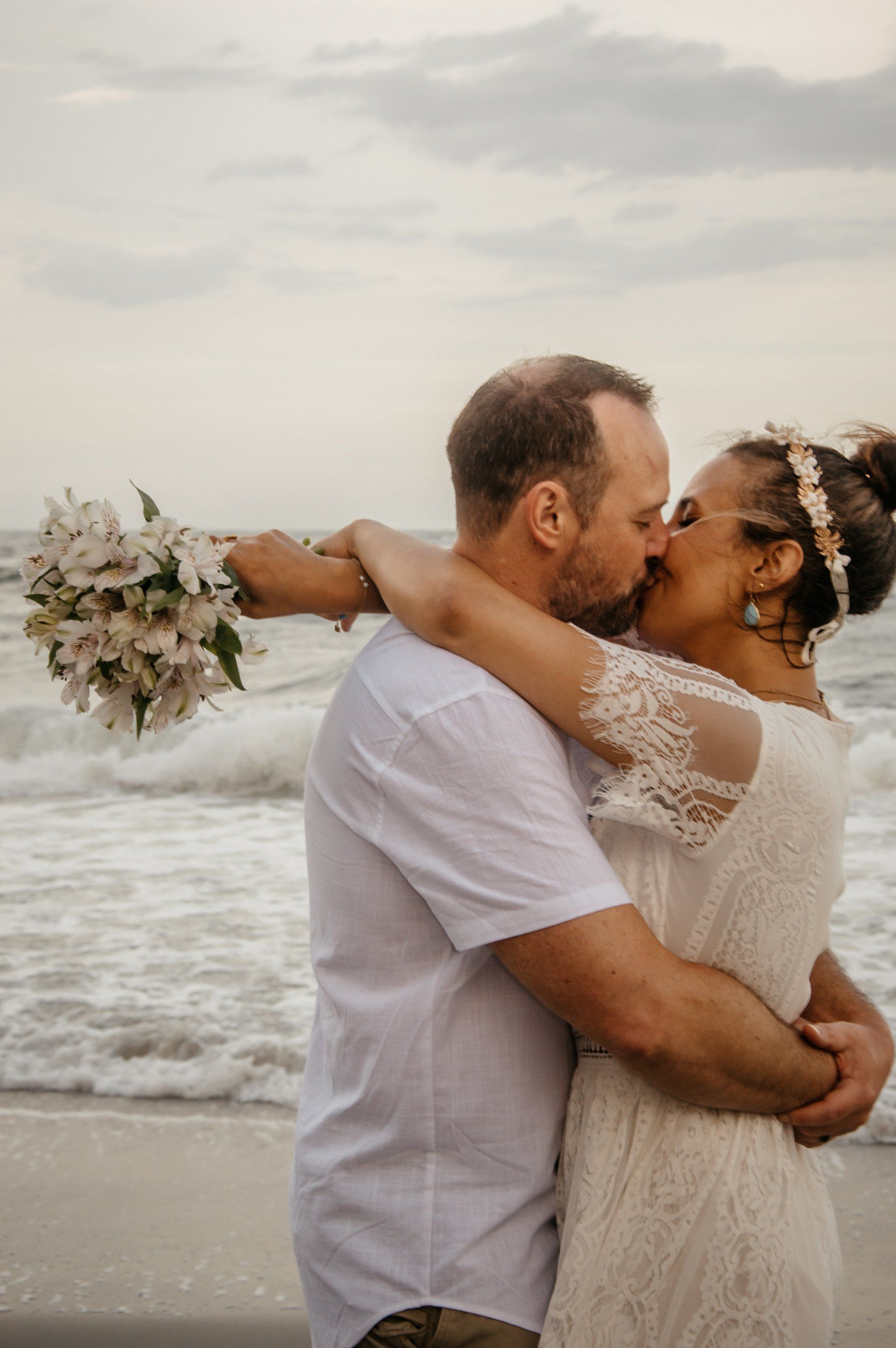 A bride and groom are kissing on the beach . the bride is holding a bouquet of flowers.