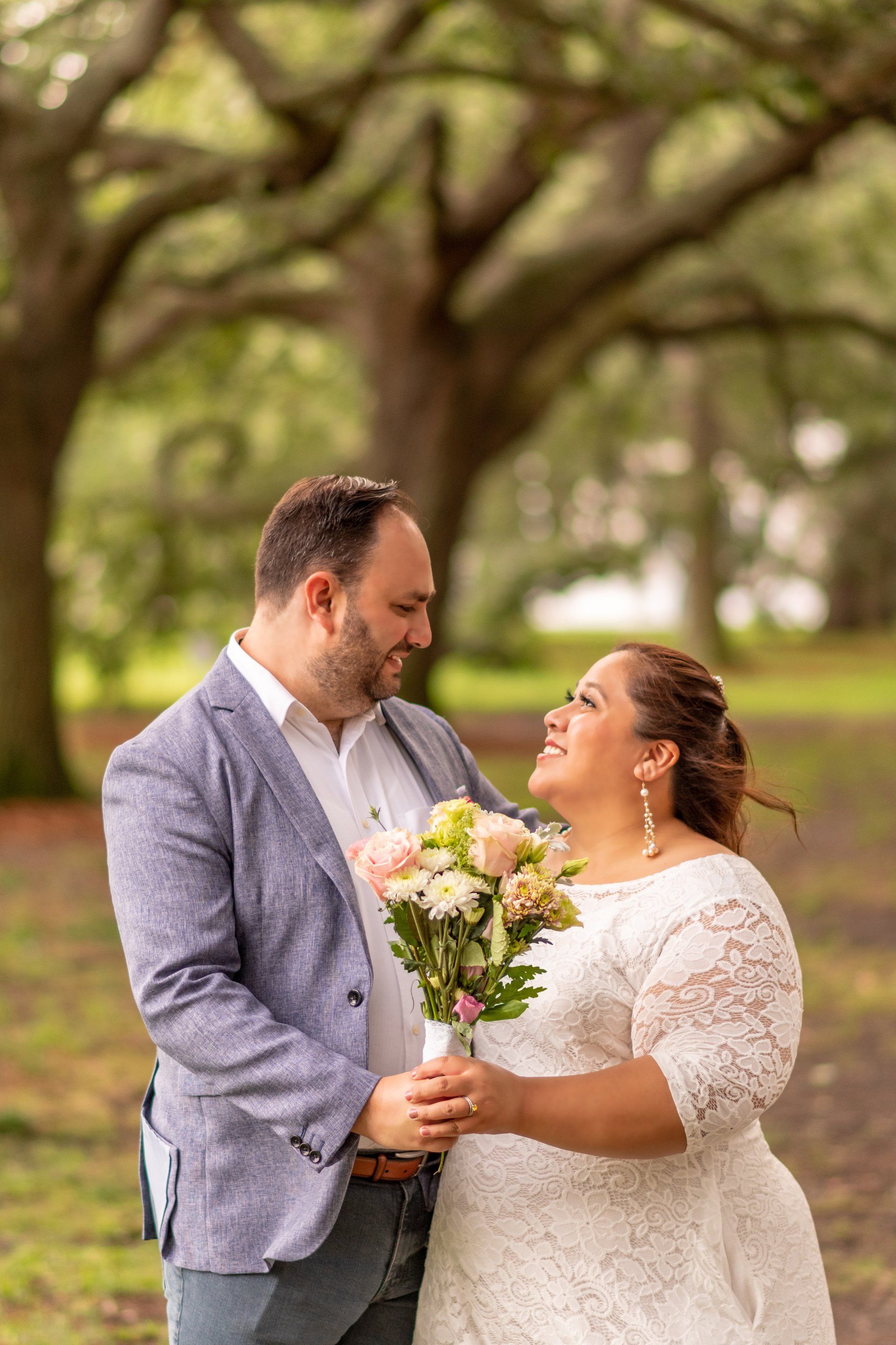 A bride and groom are standing next to each other in a park holding a bouquet of flowers.
