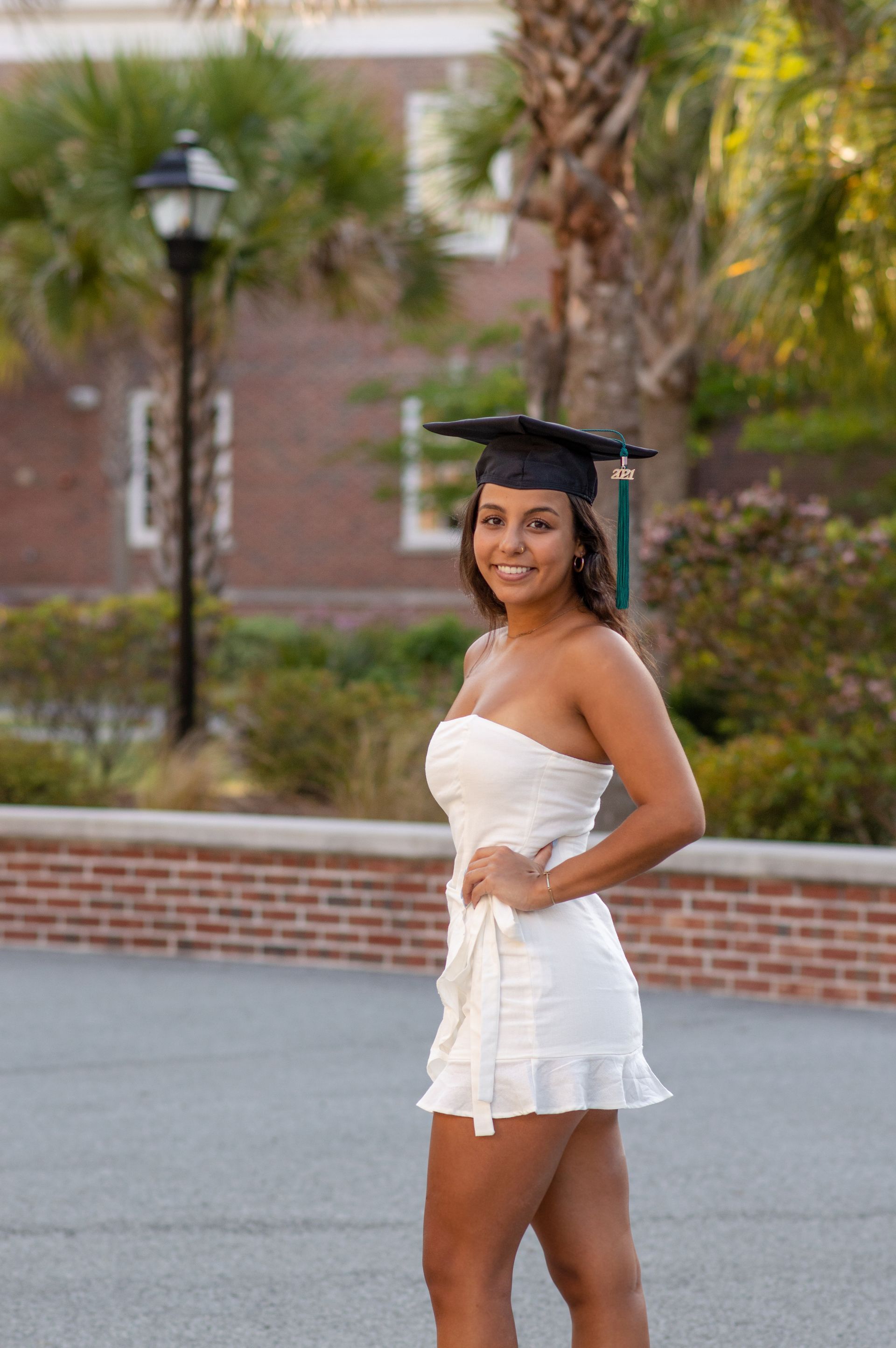 Woman in graduation cap and white dress, smiles outdoors. Brick wall and palm trees in background.