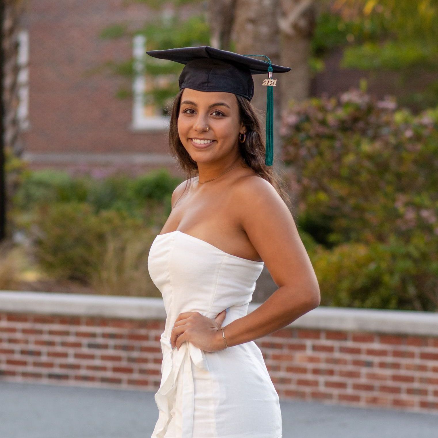 A woman in a white dress and graduation cap is standing in front of a brick wall.