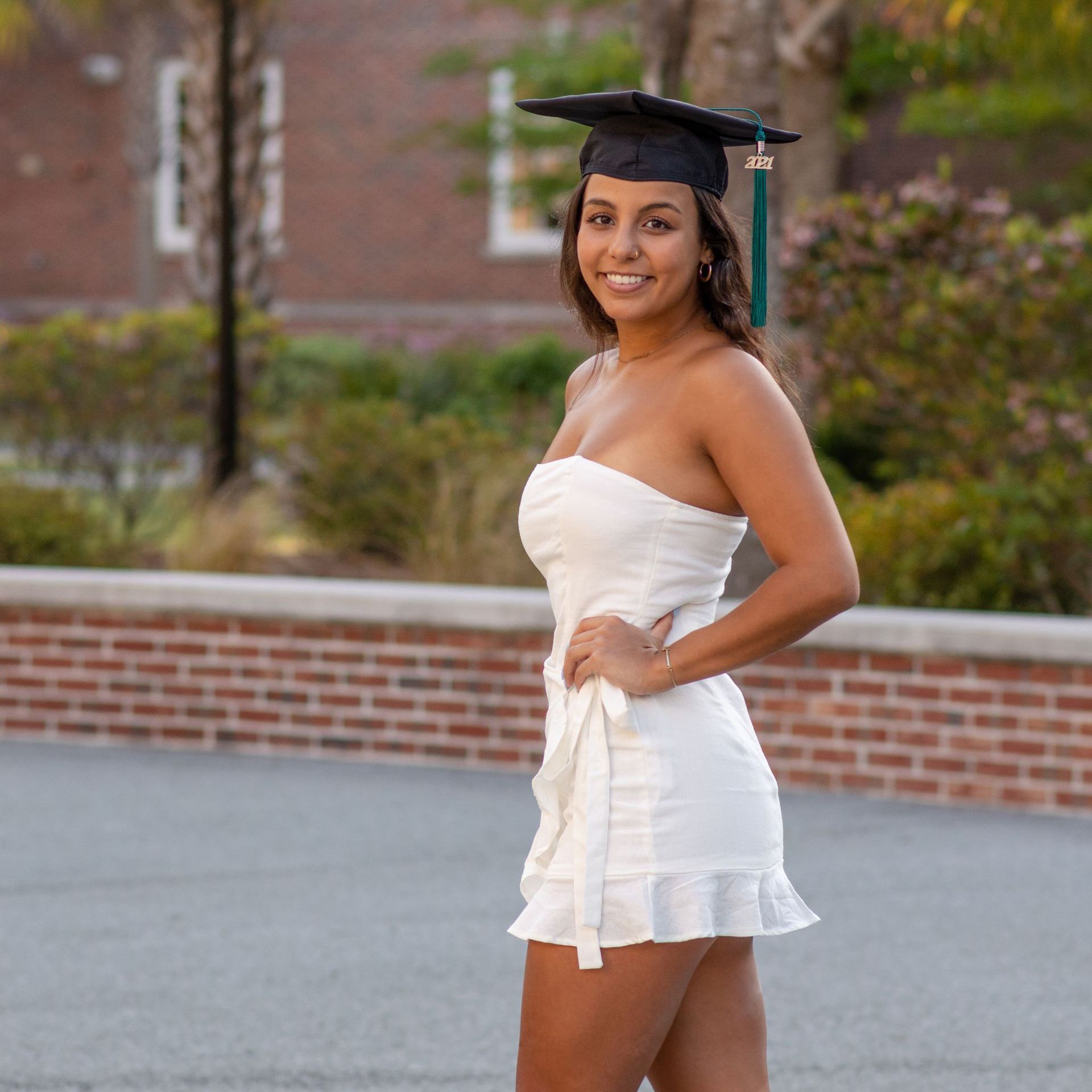 A woman in a white dress and graduation cap is standing in front of a brick wall.