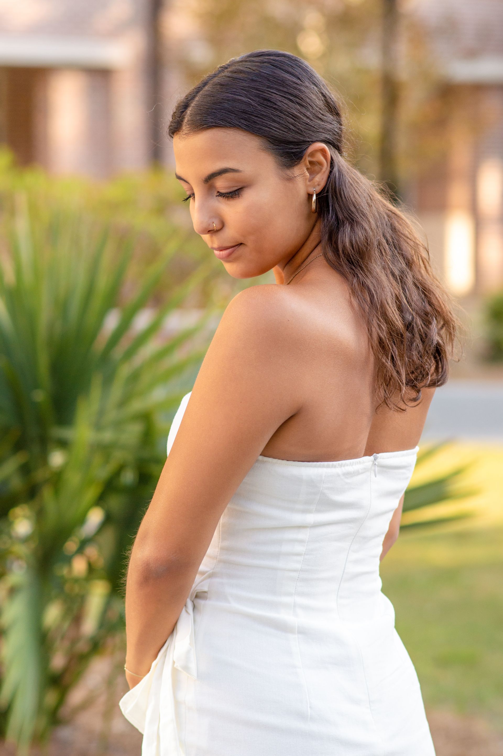 Woman in a white dress, looking down, outdoors. Brown curly hair pulled back, nose ring.