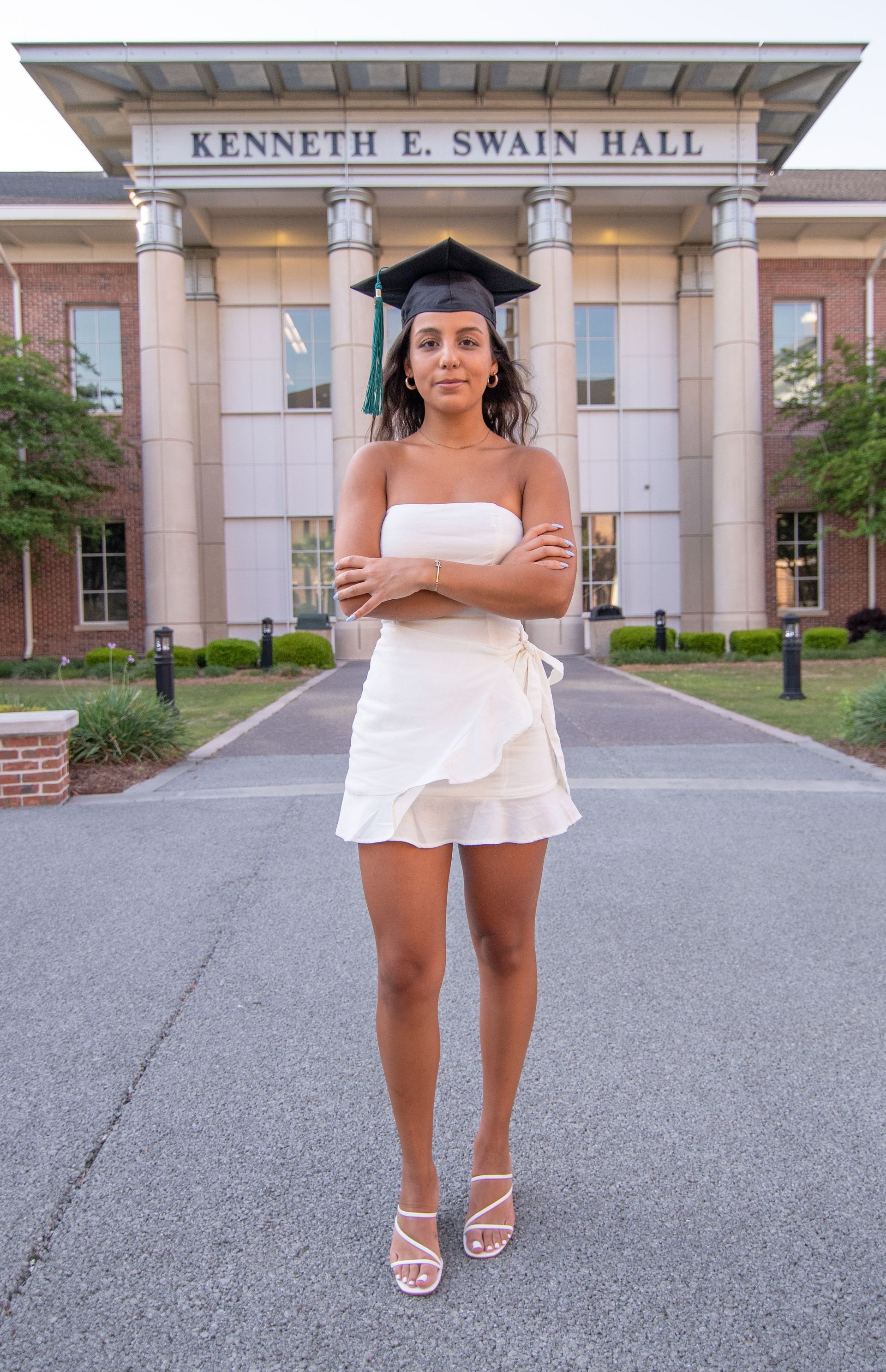 Woman in graduation cap and white dress, arms crossed, stands in front of Kenneth E. Swain Hall.