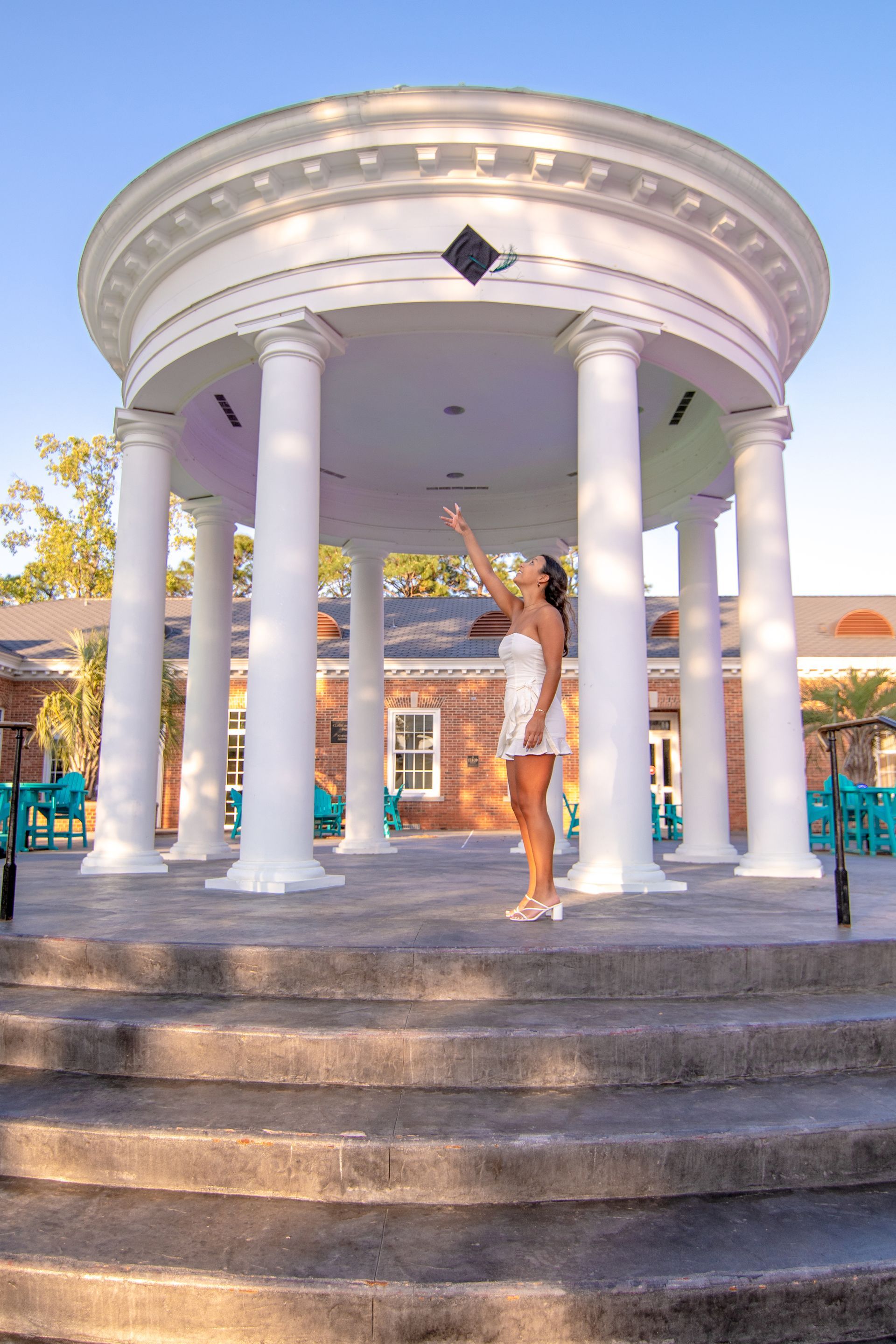 Woman in white dress points up at gazebo's center. White pillars, grey steps, and blue chairs in the background.