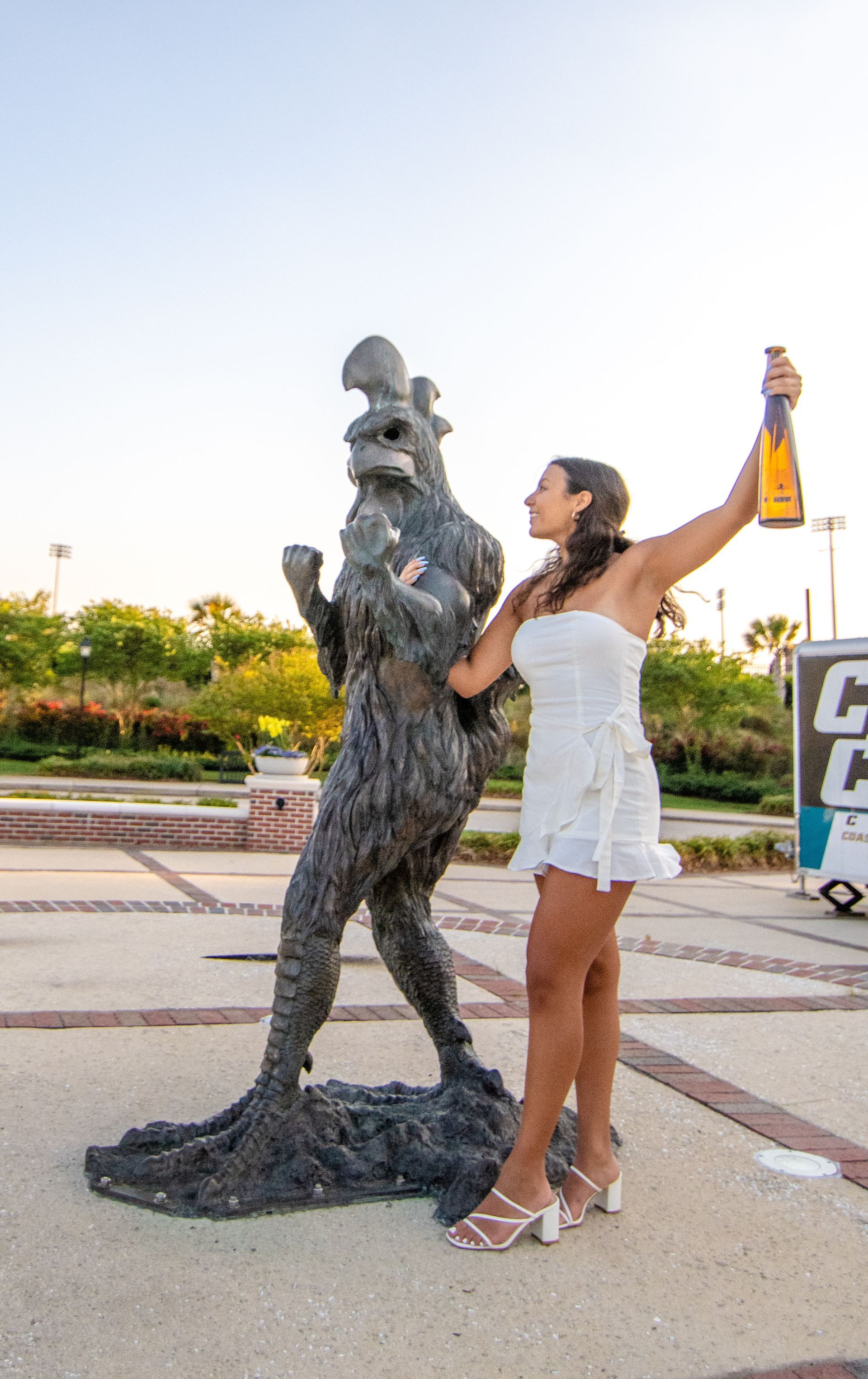 Woman in white dress, holding champagne bottle, poses with a sculpture in an outdoor park.