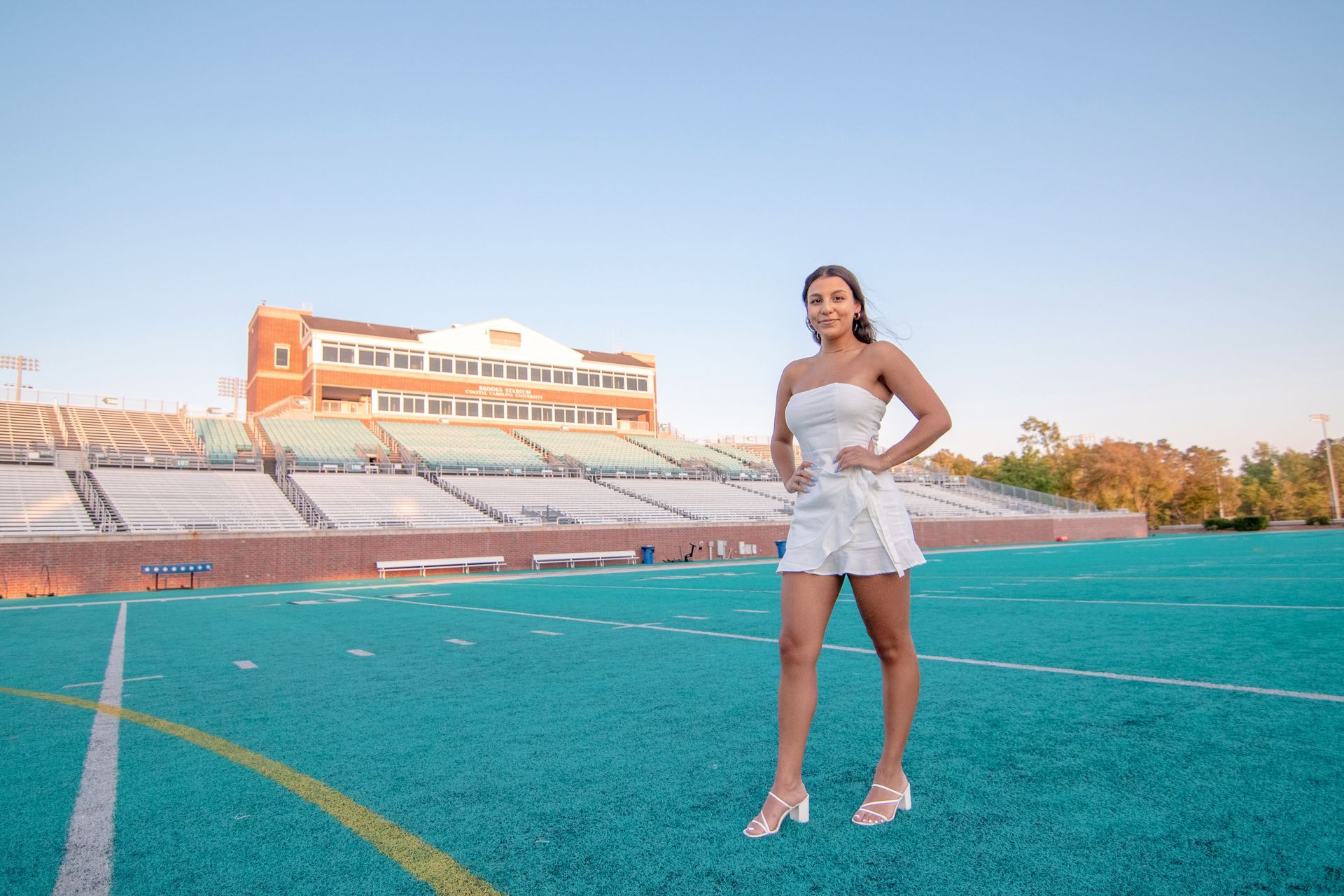 Woman in a white dress poses on a turquoise football field; stadium in background.