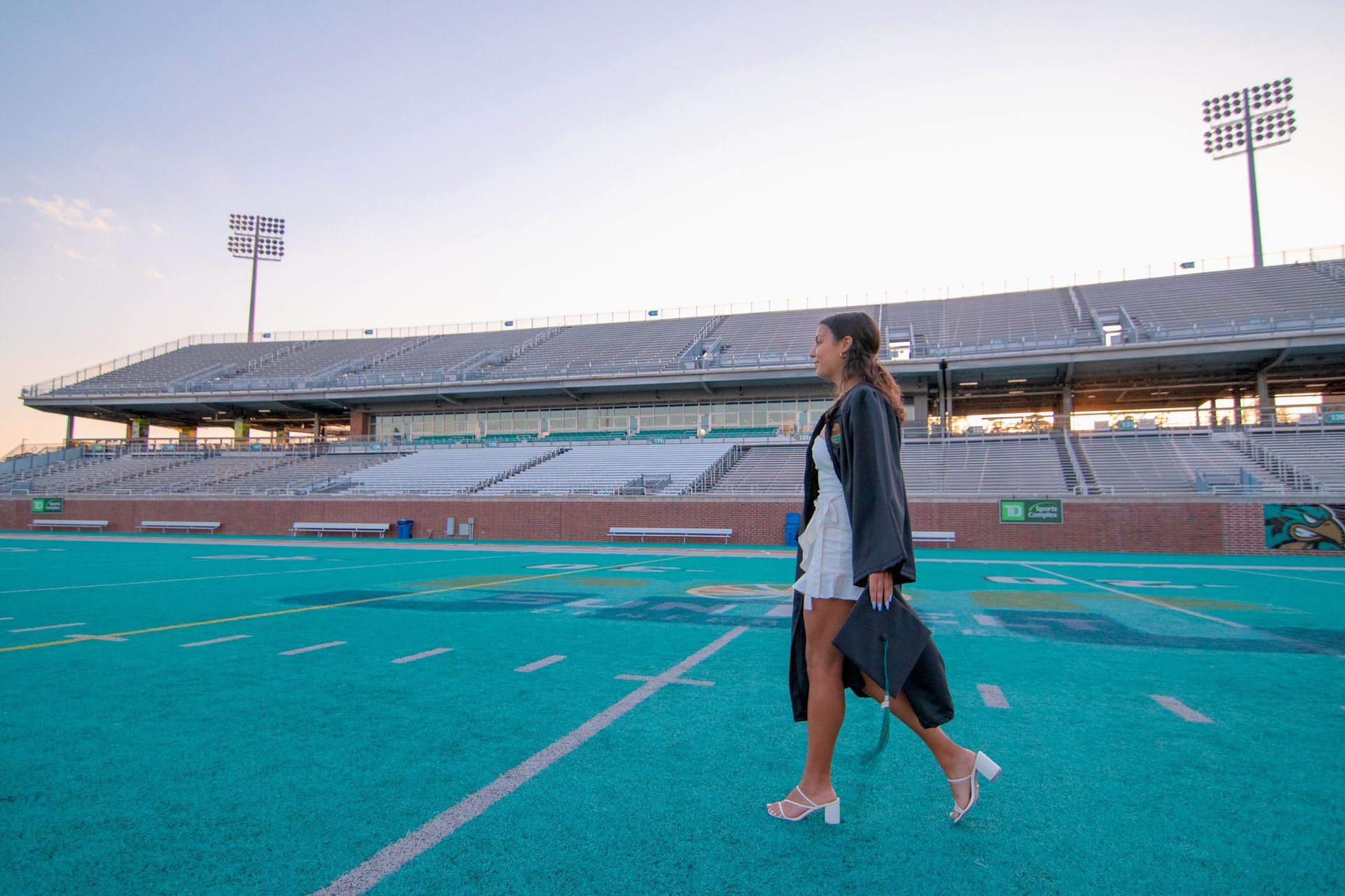 Woman in graduation gown walks on a football field. She holds cap, stadium in the background.