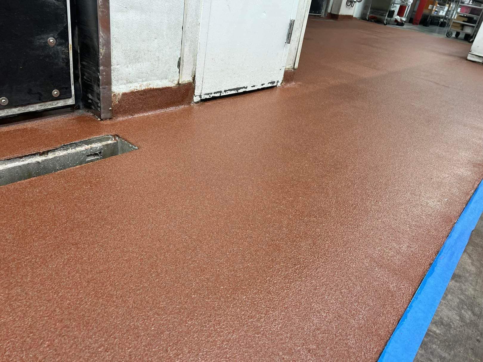 Brown speckled epoxy floor with a drain, next to a metal door, and building. Blue tape on the right edge.