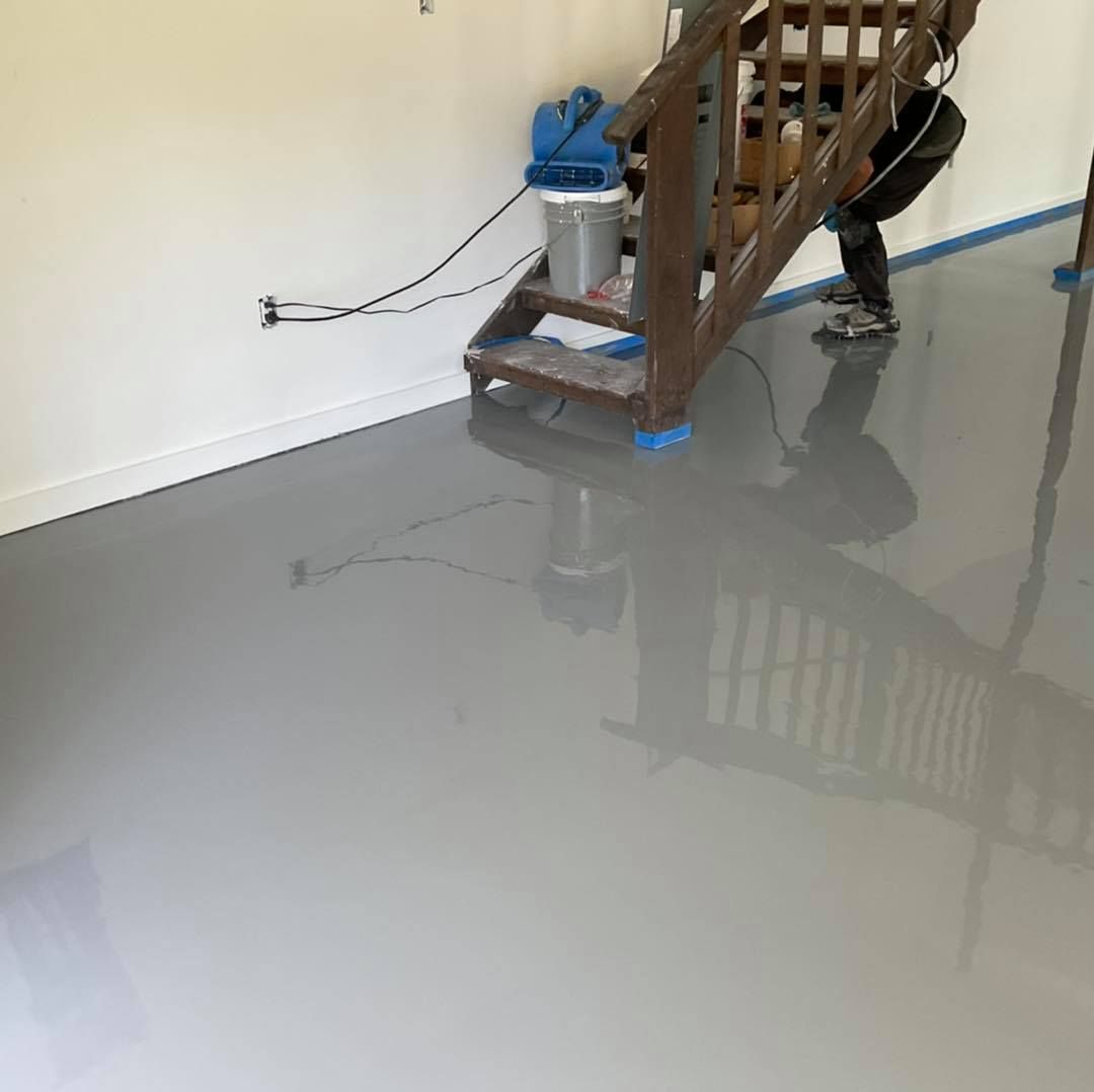 Person applying gray epoxy to a garage floor. Stairs and equipment visible; blue tape at baseboards.