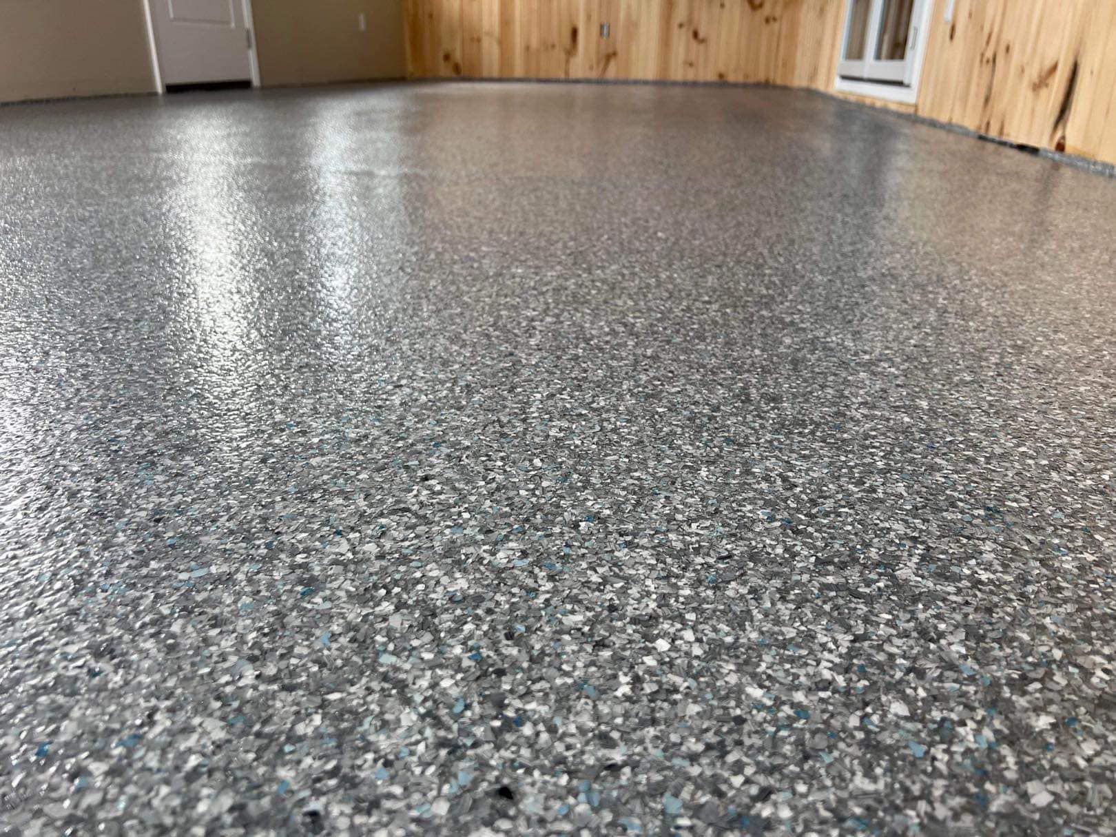 Close-up of a shiny, speckled epoxy floor with gray and white chips. Wooden paneling is visible along the walls.