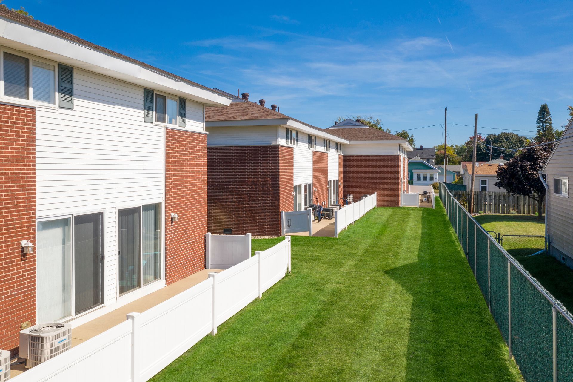 Row of brick and white townhouses with a green lawn and white fence. Clear blue sky.