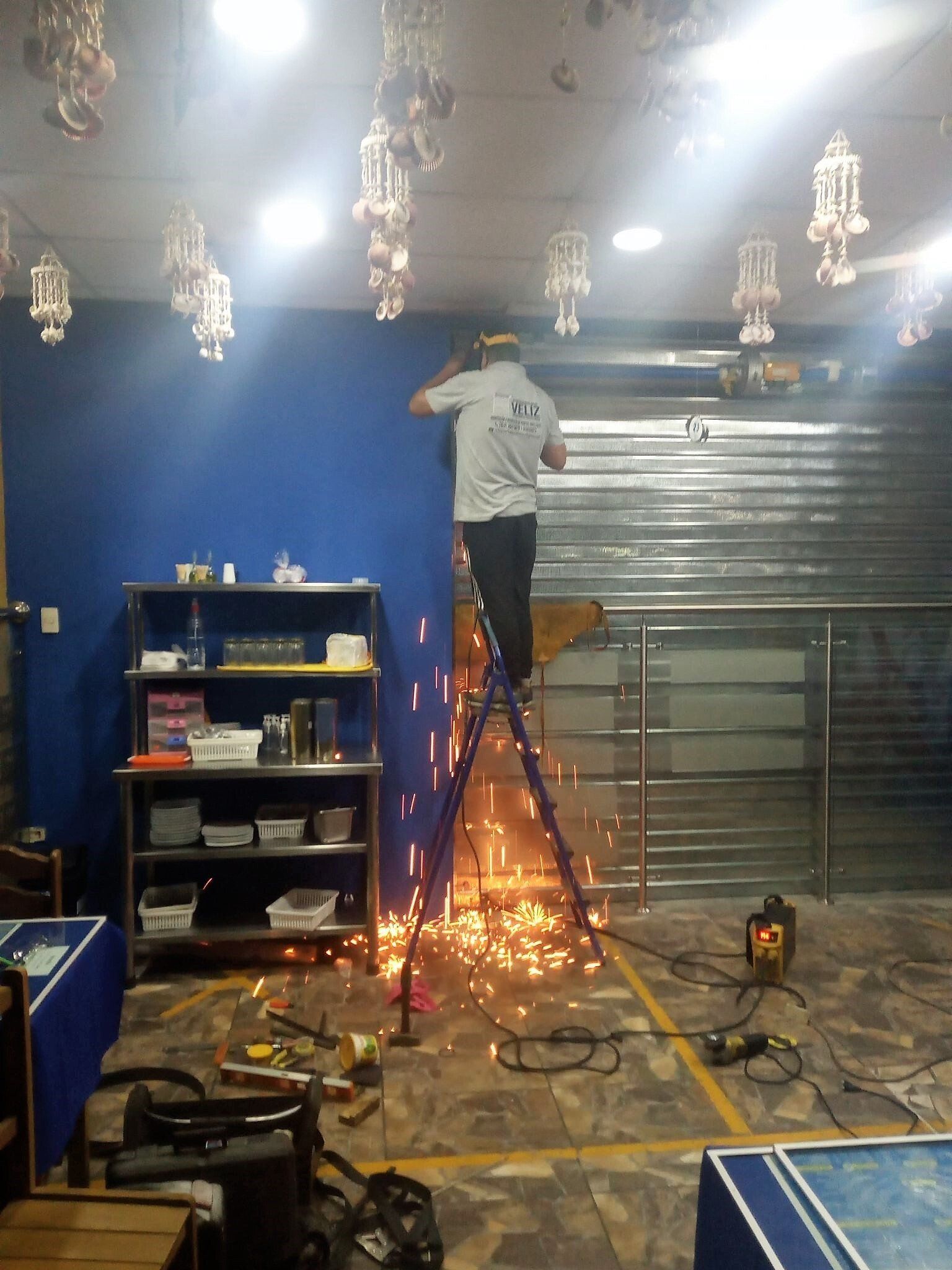 Una persona subida a una escalera trabajando en las luces del techo de una habitación azul, con chispas que caen del piso de abajo.