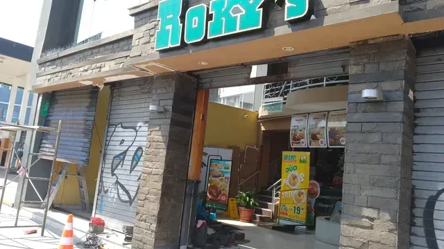 Local comercial con letrero color turquesa, entrada abierta y estanterías llenas de productos en el interior, ubicado en una calle soleada.