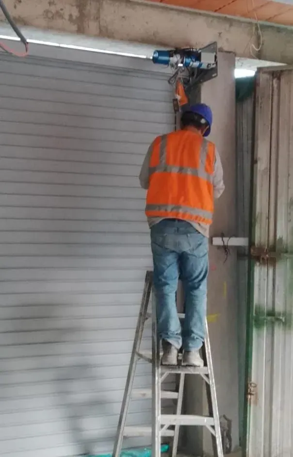 Un trabajador con chaleco de seguridad naranja, subido a una escalera, ajusta el motor de una puerta de garaje sobre una persiana cerrada.