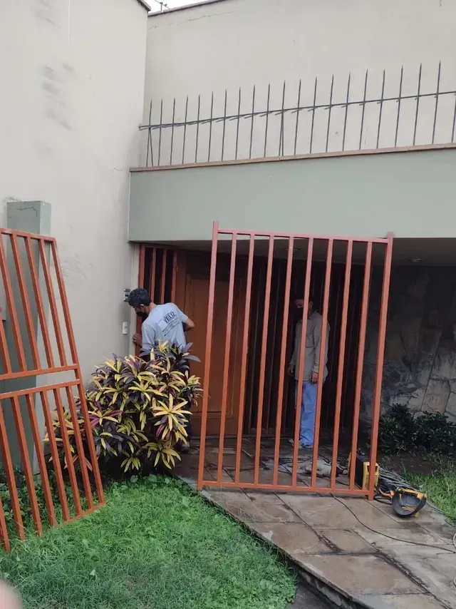 Dos personas mueven una gran puerta metálica rosa en el exterior de una casa con un pequeño césped y un camino de acceso.
