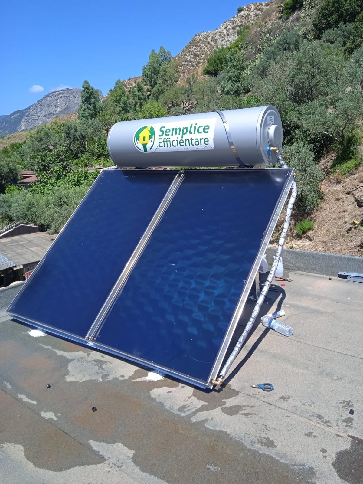 Scaldabagno solare su un tetto con pannelli blu e un serbatoio argentato, sullo sfondo colline e cielo azzurro.