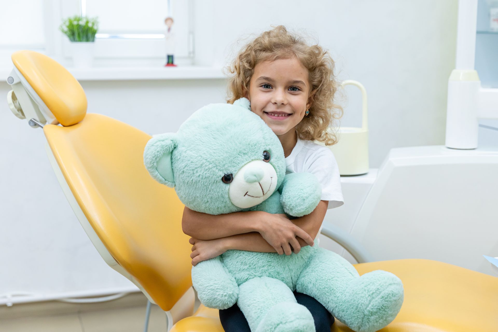 Young girl with curly hair smiles, hugging a teal teddy bear in a dentist's chair.