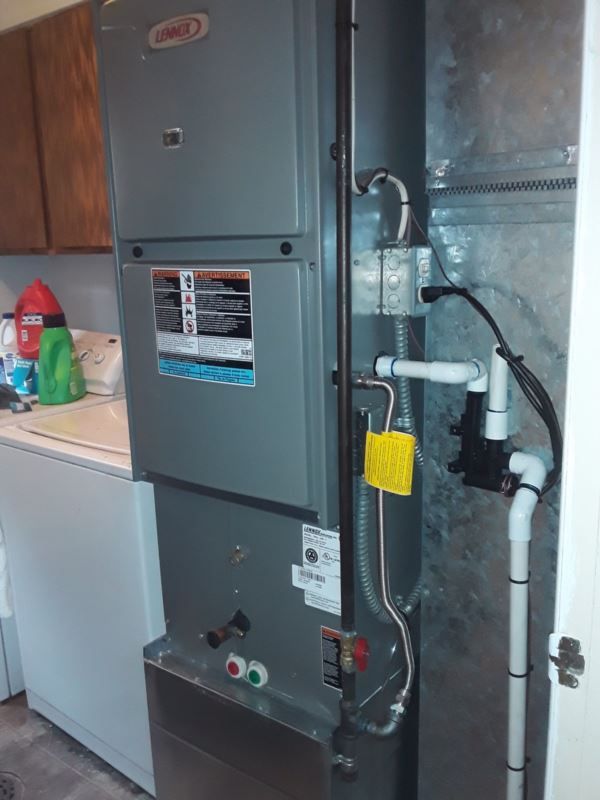 Gray Lennox furnace next to a metallic duct, in a laundry room. White pipes and yellow tag are visible.