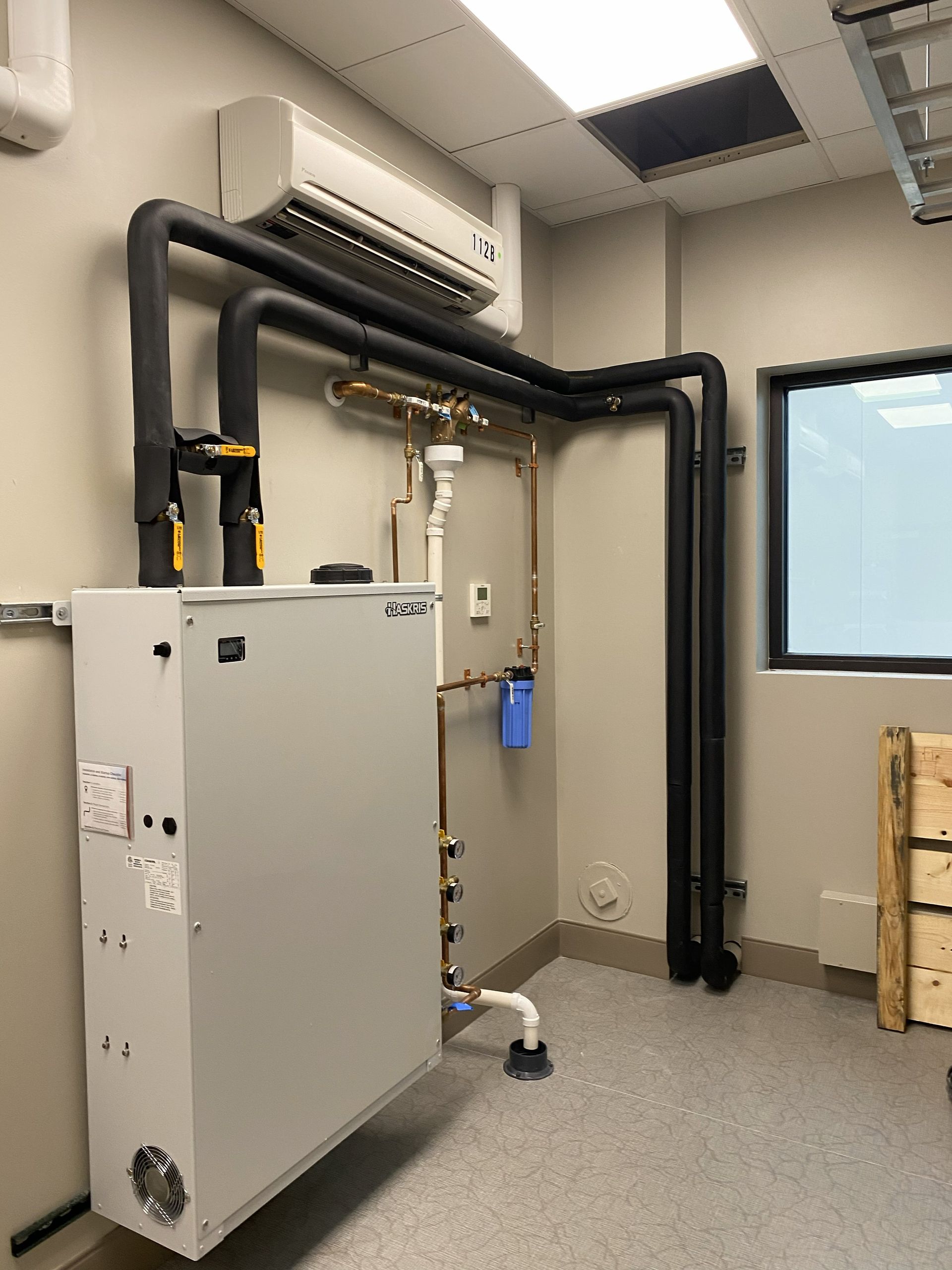 HVAC system with a grey unit, black pipes, and a white air conditioner on a wall in an indoor setting.