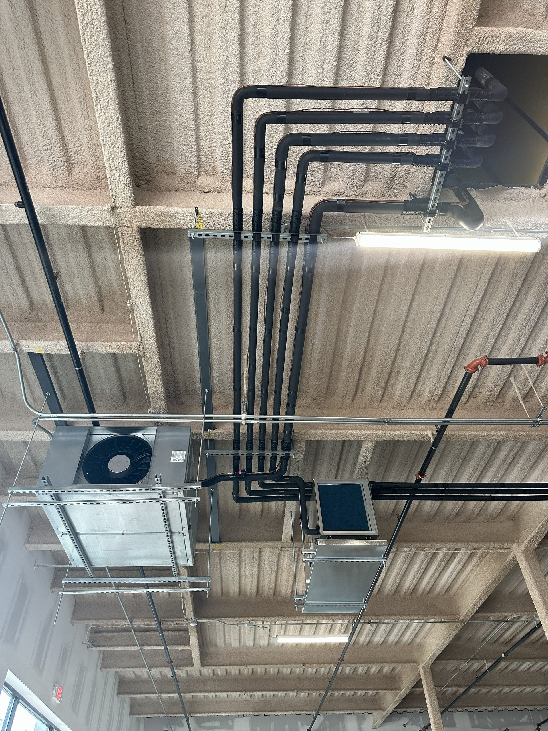 HVAC ductwork and piping on a concrete ceiling, black insulated pipes, metal air handlers, and a fluorescent light.