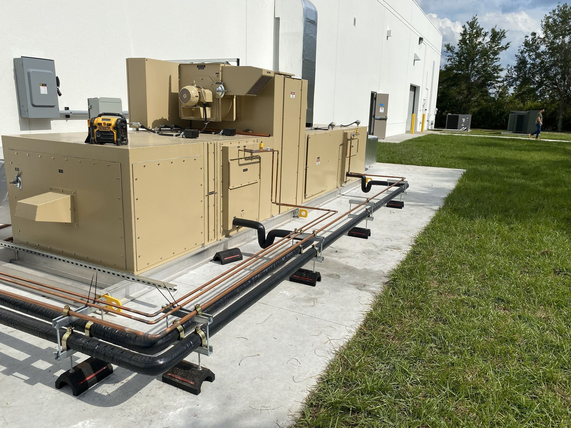 Large beige HVAC unit on concrete pad next to a white building and grass. Piping runs along the ground.