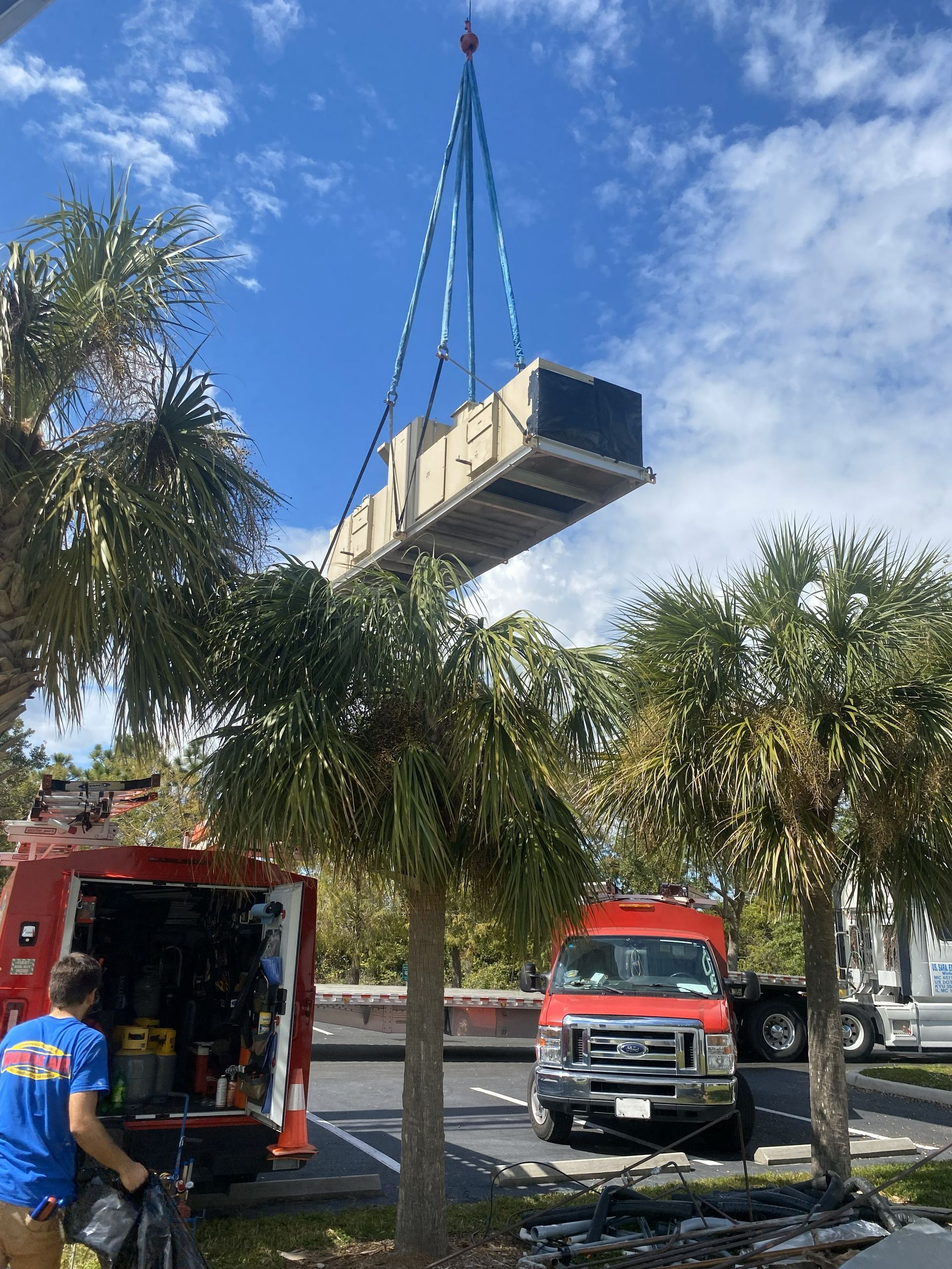 Crane lifting rectangular HVAC unit near palm trees and a work truck.