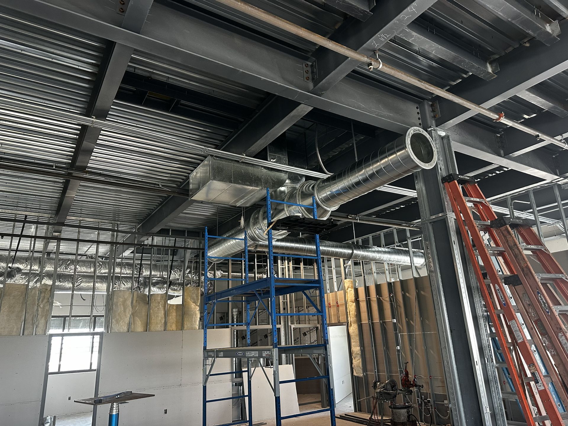 Construction site interior with exposed ductwork, metal beams, scaffolding, and ladder.