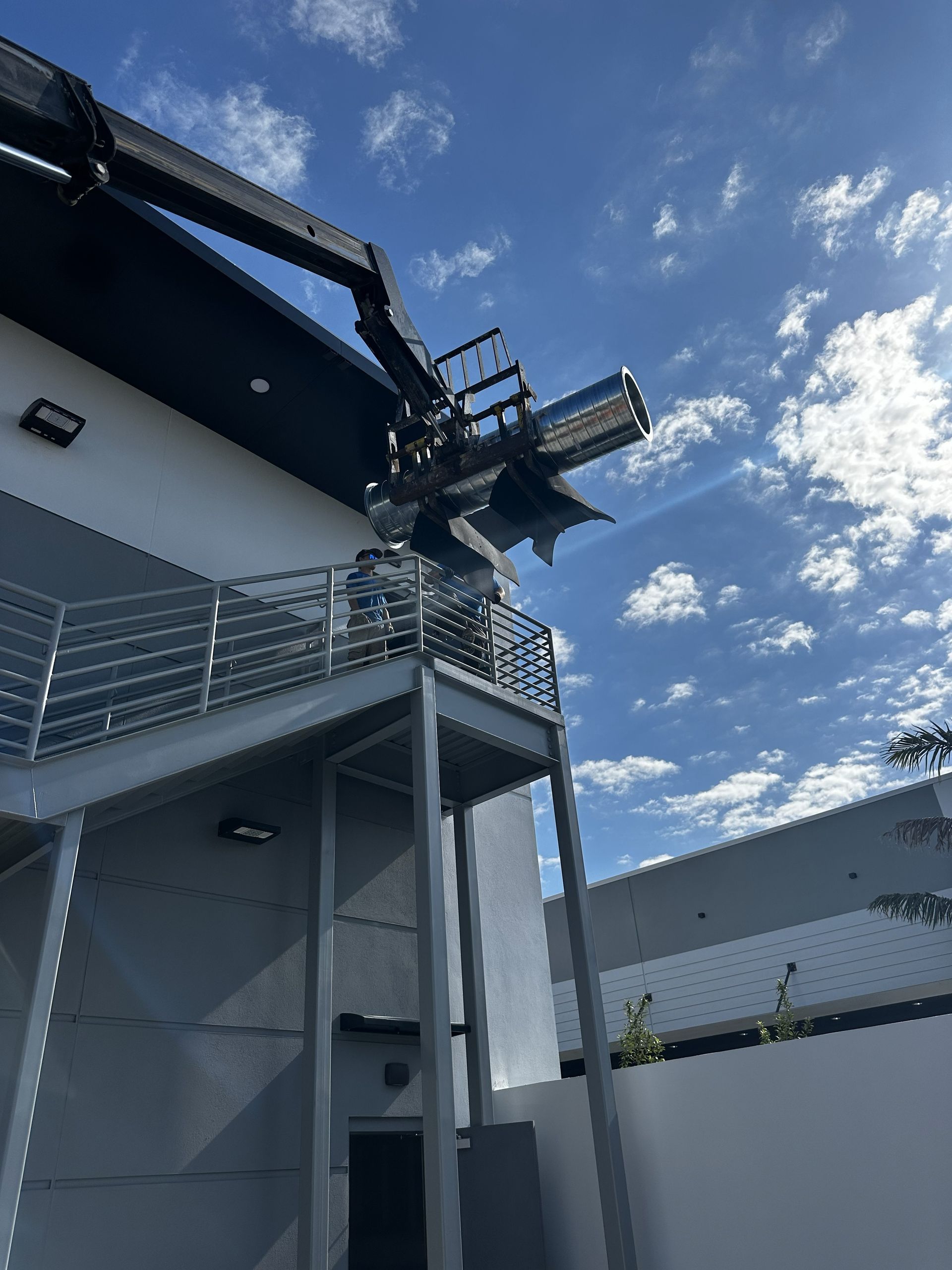 Crane lifting a silver object onto a building's exterior. Blue sky with clouds; metal staircase on the building.