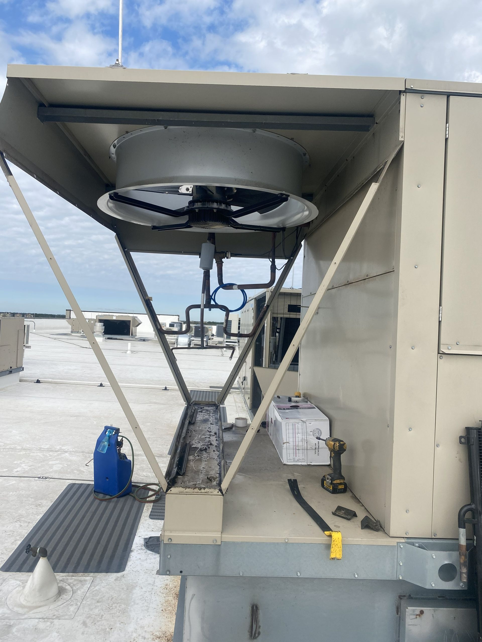 HVAC unit on a rooftop with fan, supports, and tools. Beige and gray colors, under a cloudy sky.
