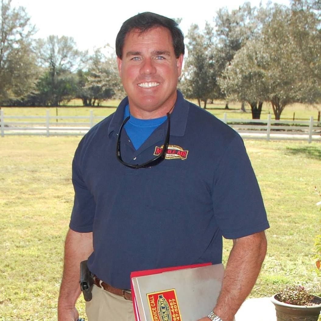 Man in blue polo shirt holding a binder, outdoors in front of a white fence and trees.