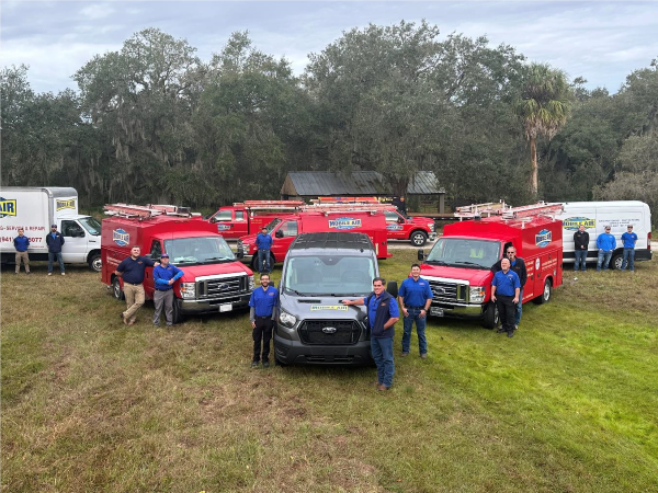 Group of men pose with work vans on a grassy field. Red, white, and gray vans, plus tall trees.