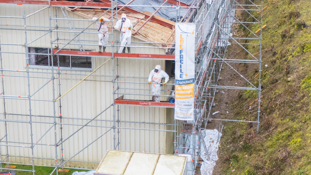Toit en ardoise grise percé de deux puits de lumière reflétant un ciel bleu et le bâtiment adjacent.