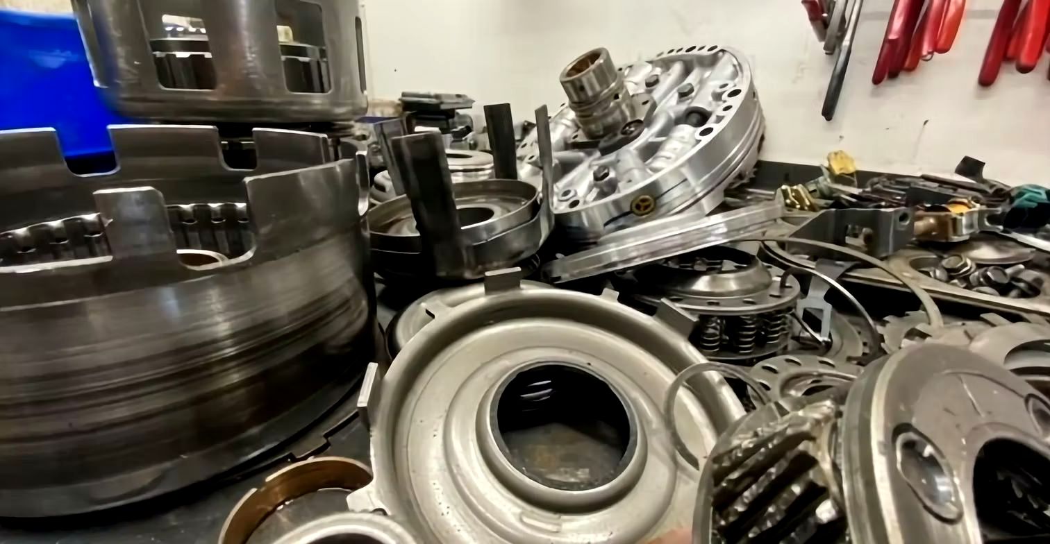 Disassembled car transmission components on a workbench. Metal parts, gears, and housings are scattered, likely for repair.