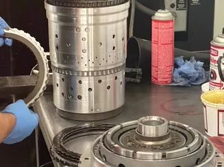 A person in blue gloves assembling automotive transmission parts on a worktable. Cylindrical metal components are visible, along with a spray can and lubricant.