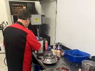 Mechanic in red and black uniform working on car parts in a repair shop. A transmission sits on the workbench.