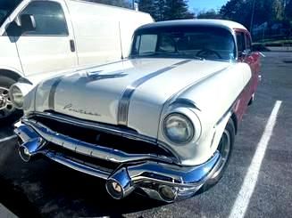 White and maroon 1950s Pontiac car parked next to a white van in a parking lot. Chrome bumper and trim.