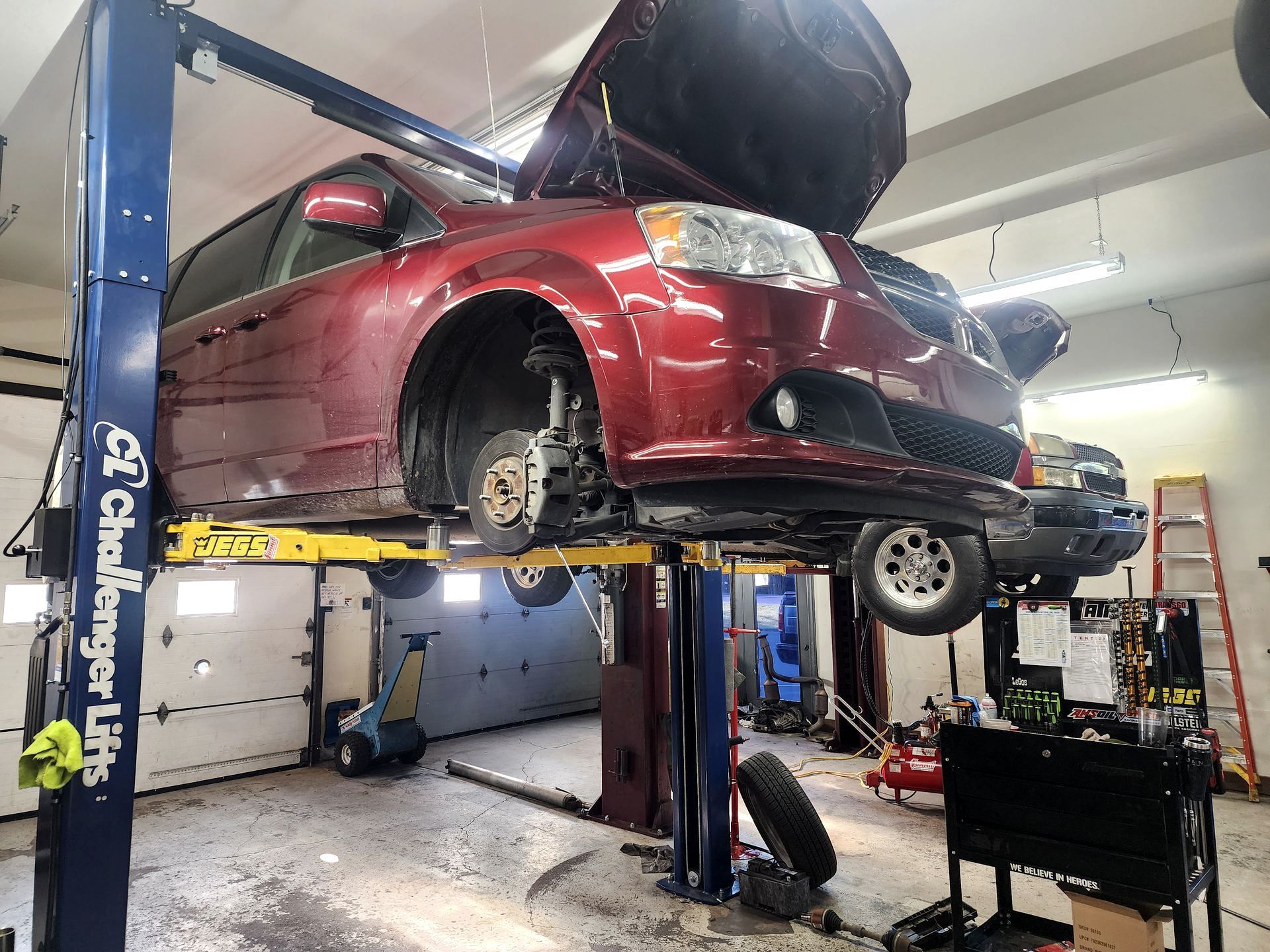 A red minivan on a lift in an auto repair shop. The hood is open, and a tire is removed.