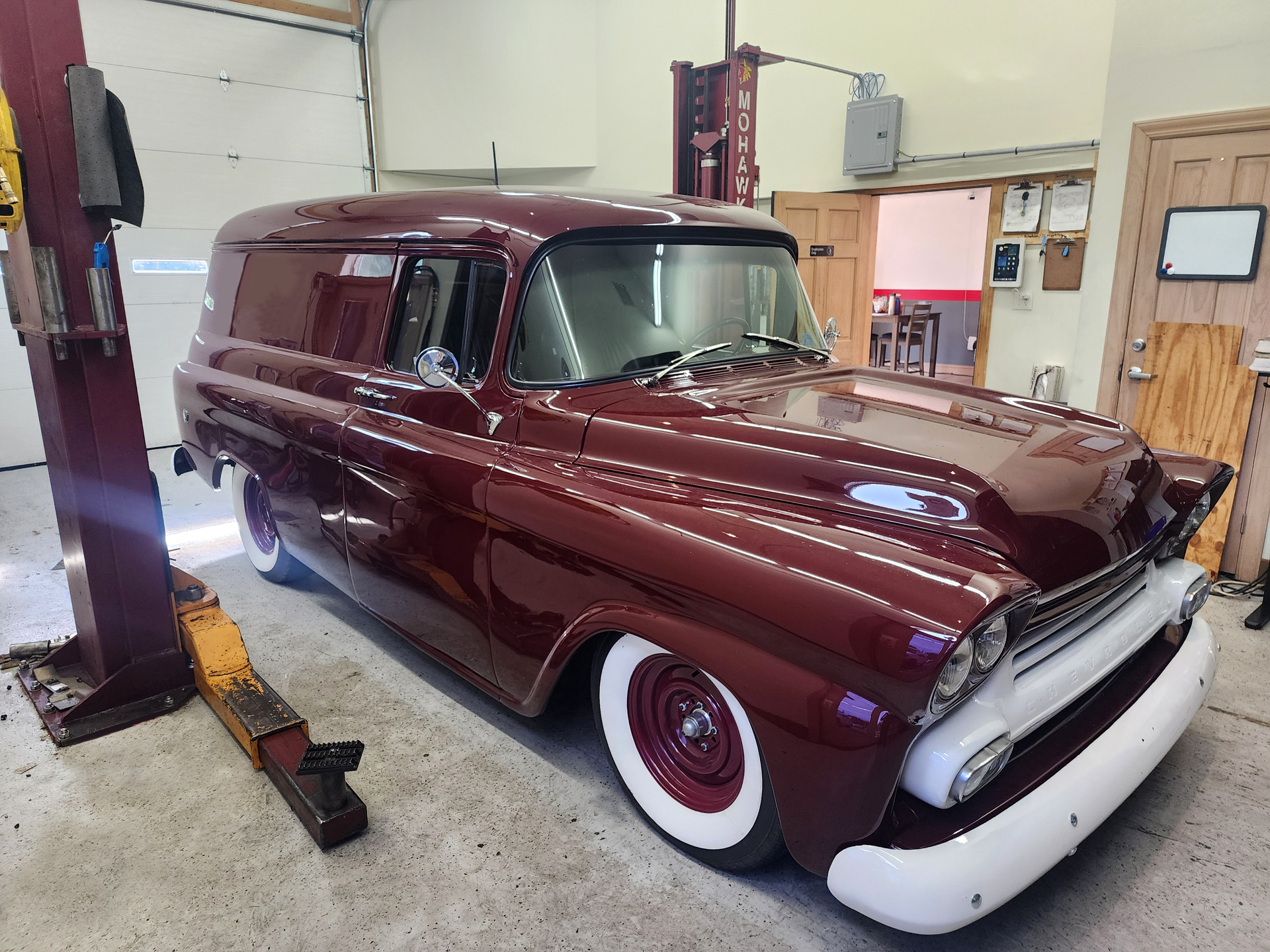 Maroon custom vintage panel van in a garage, parked next to a lift. White wall tires and white accents.