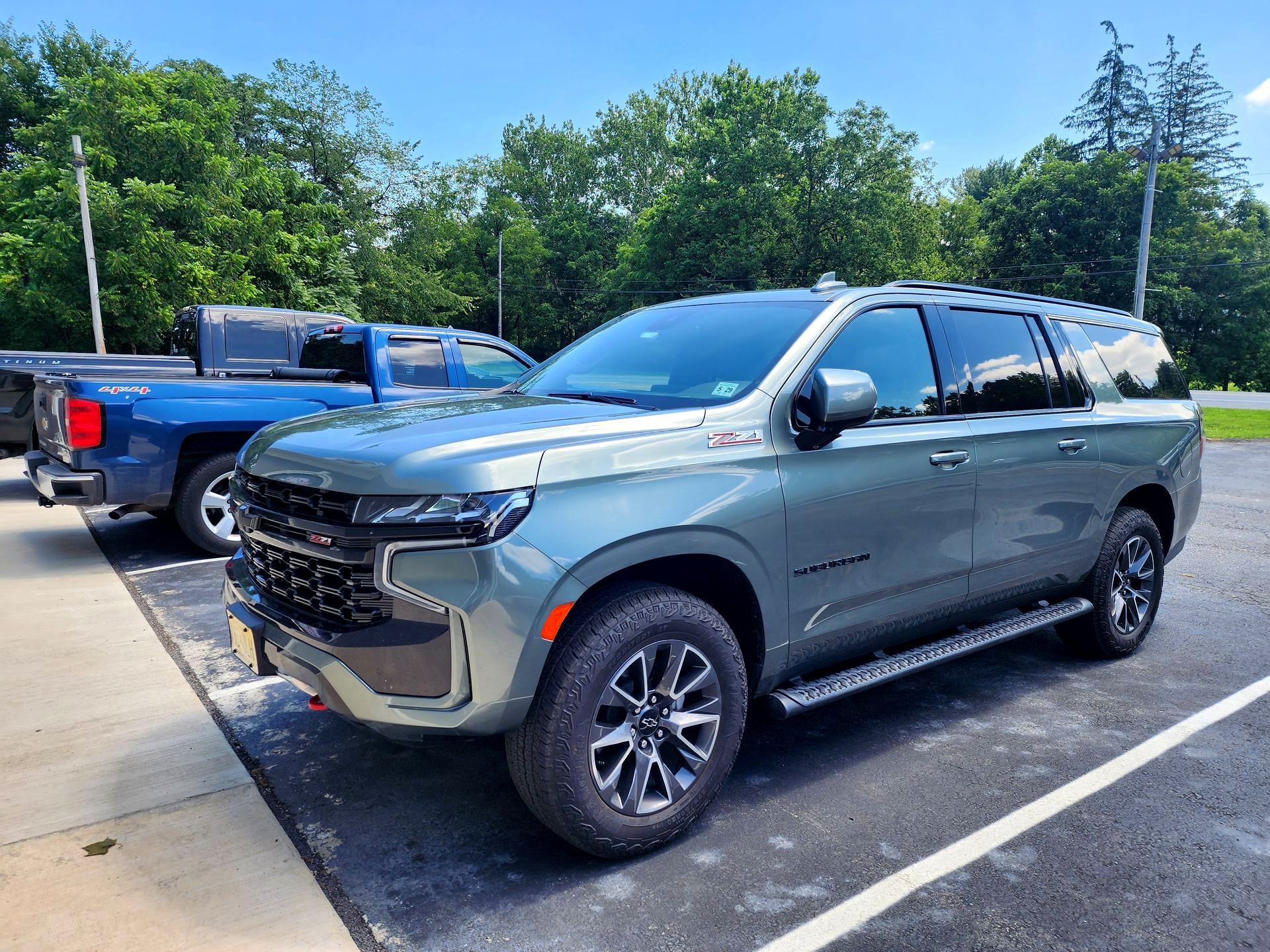 Green Chevrolet Suburban parked outdoors on a sunny day. A blue pickup truck is visible behind it.