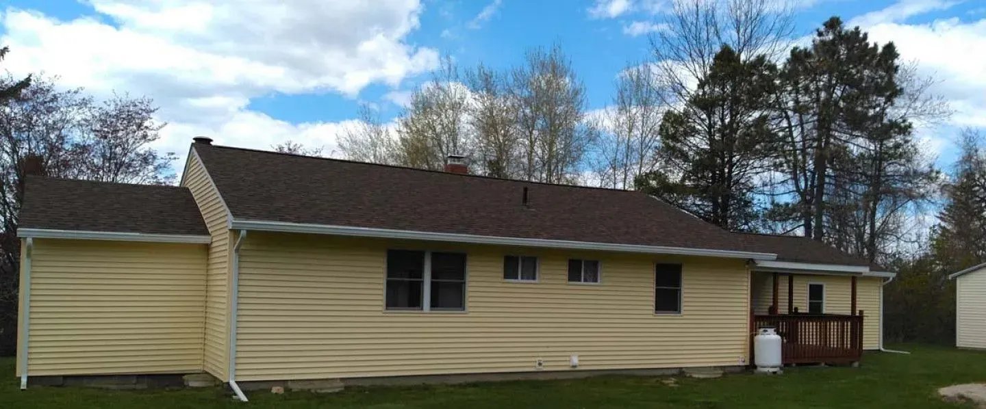 A yellow house with a brown roof is surrounded by trees and grass.