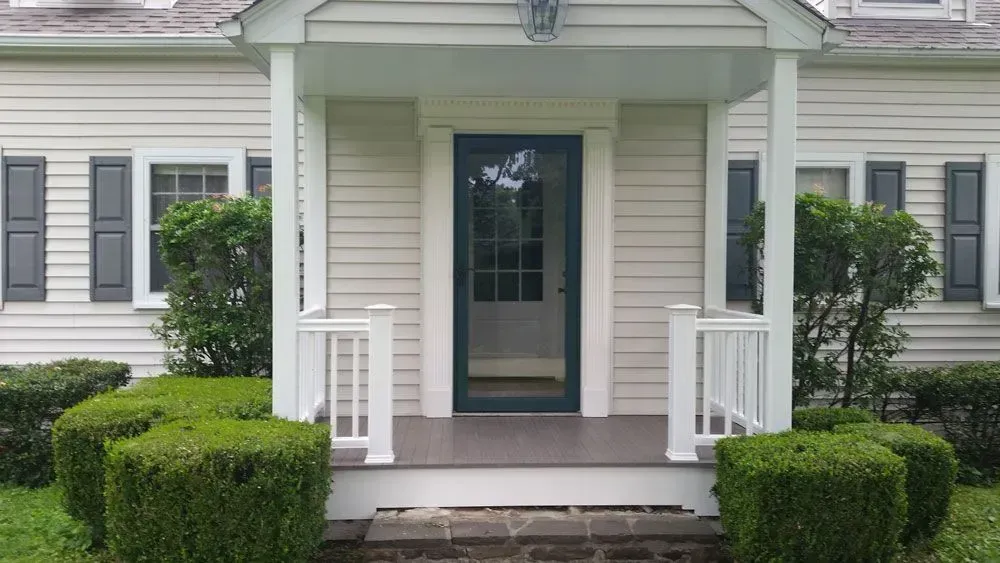 A white house with a porch and bushes in front of it.