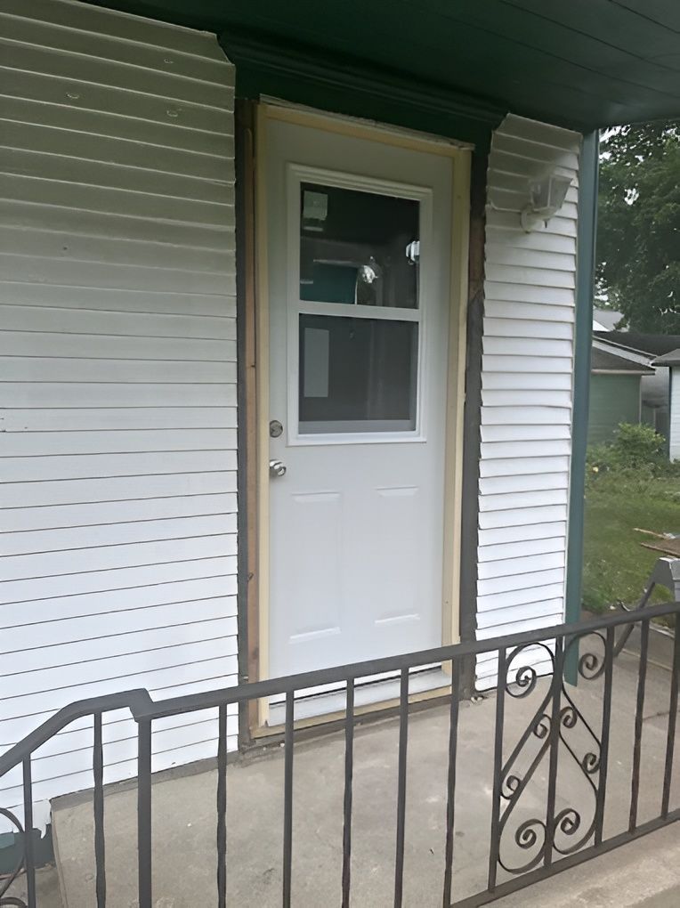White door on a porch with decorative iron railing, siding on the house, and green trim.