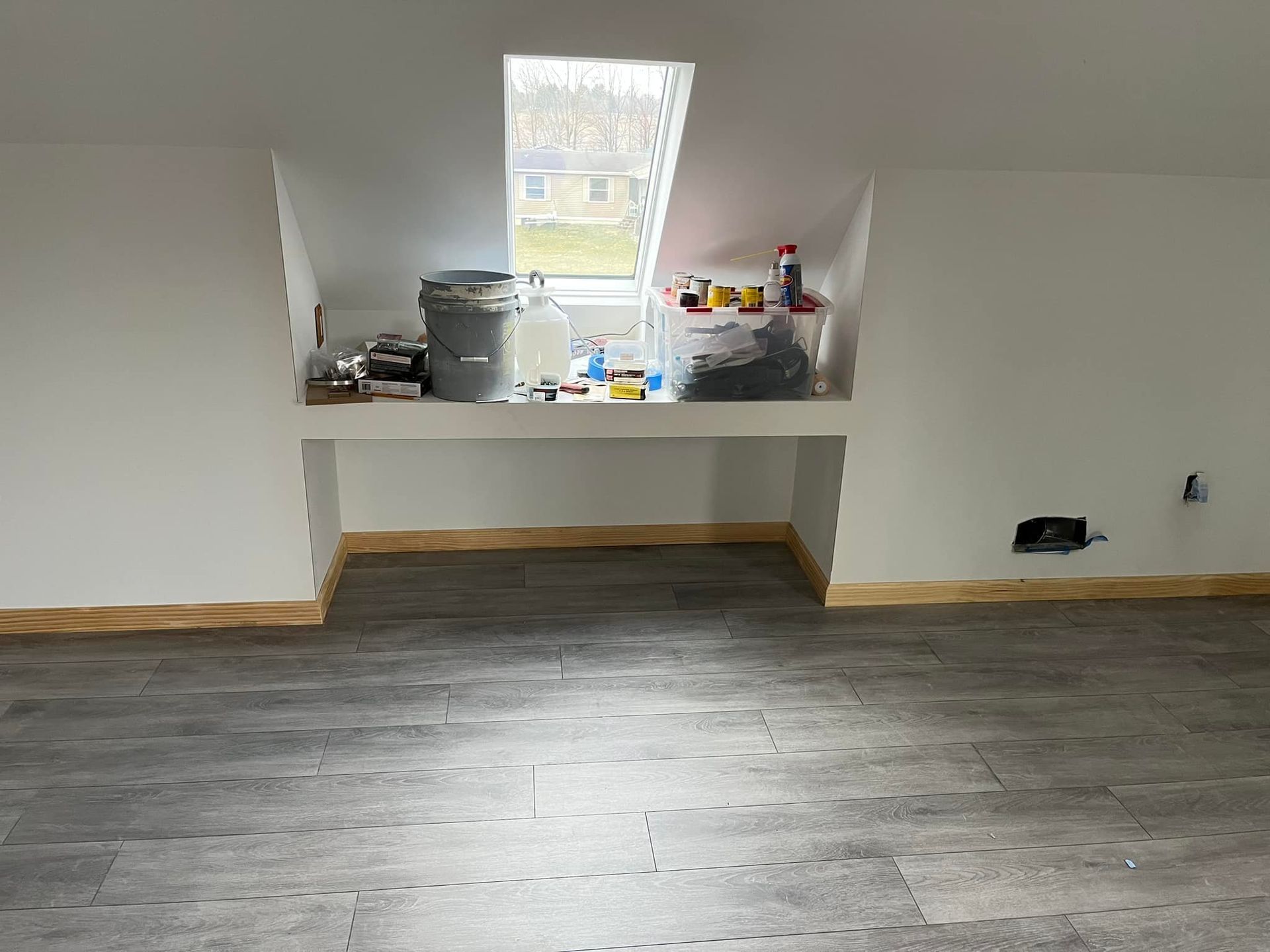 Attic room with a built-in shelf under a window, containing various items. Gray flooring and white walls.