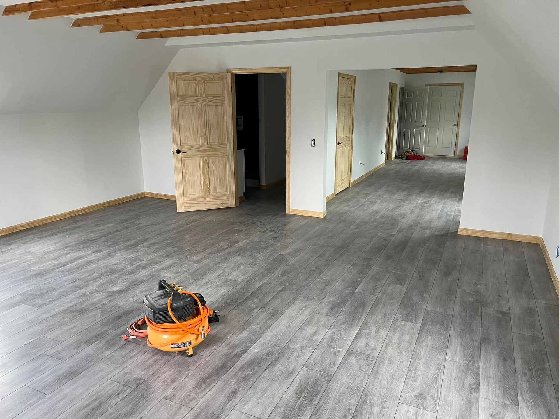 A room with gray vinyl flooring, unfinished wood doors and ceiling beams; an orange air compressor rests on the floor.